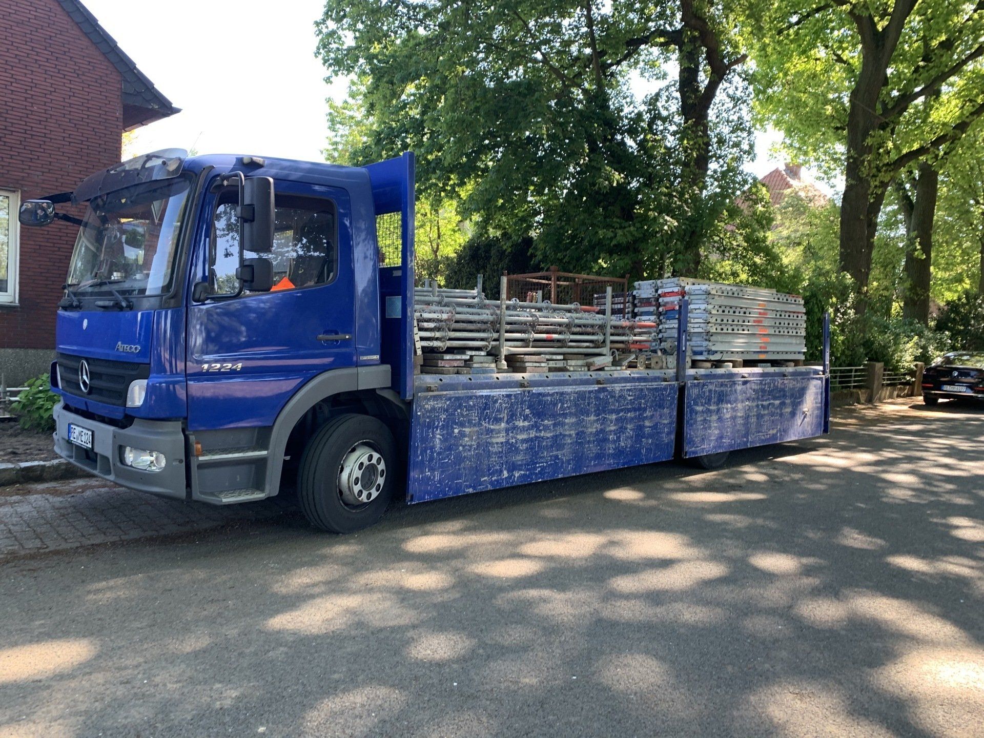 Blauer Arbeitslastwagen mit Gerüsten auf einer Straße geparkt. Gebäude und Bäume im Hintergrund.