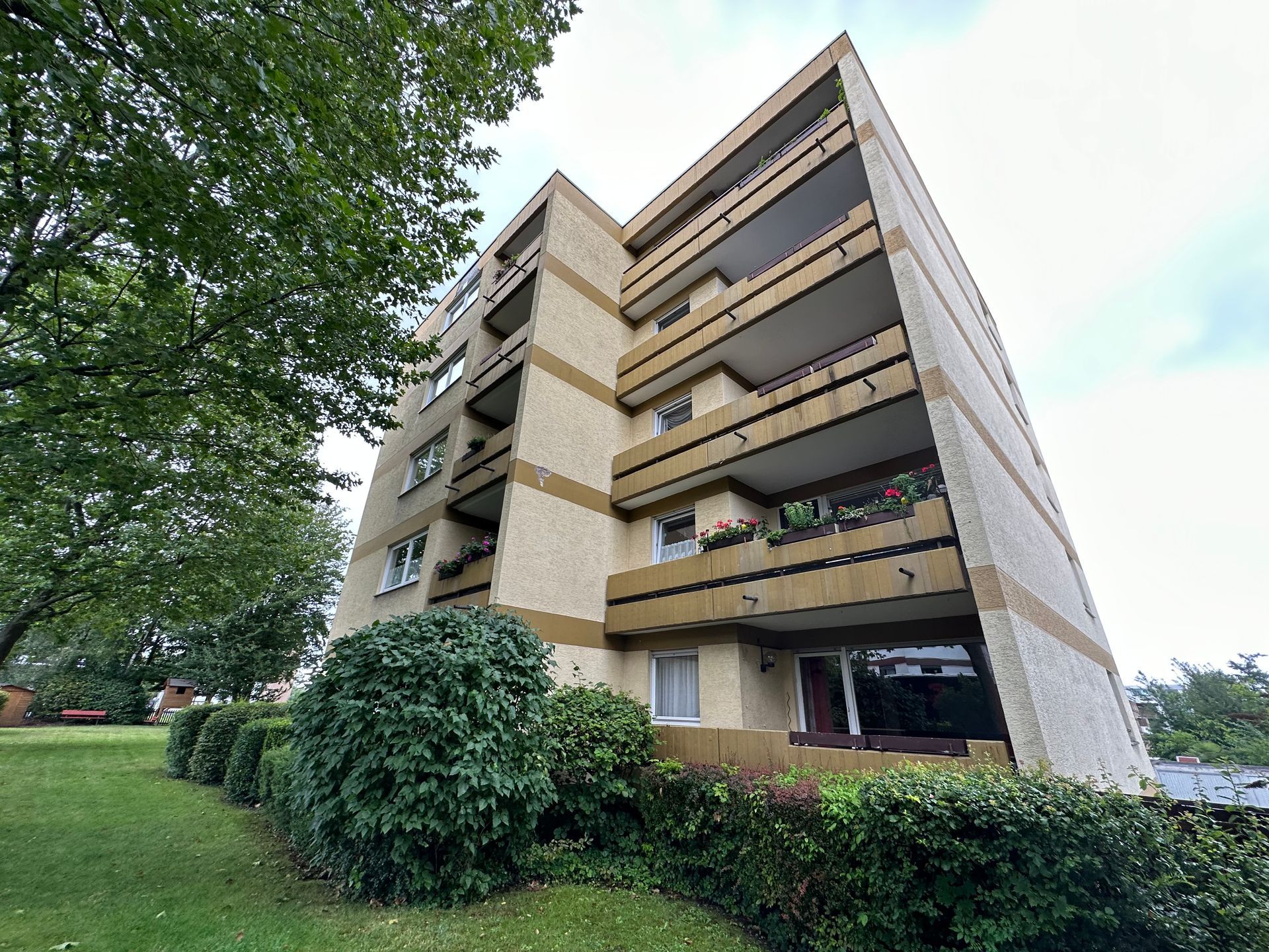 Mehrstöckiges Wohnhaus mit Balkonen, beiger Fassade, grünen Büschen und bewölktem Himmel.