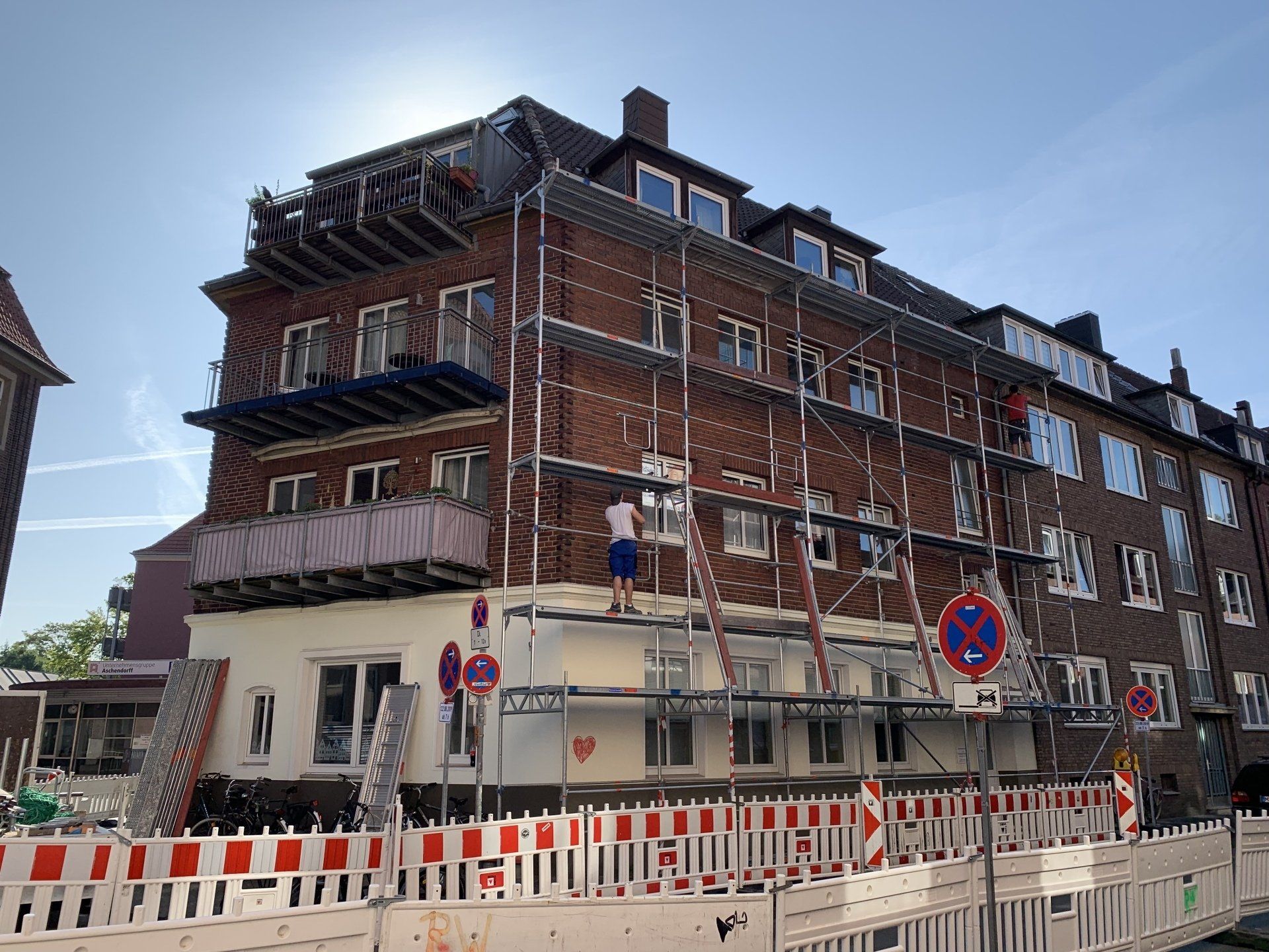 Gebäude im Bau mit Gerüst, Arbeitern, Ziegelfassade, weiß-roter Absperrung.