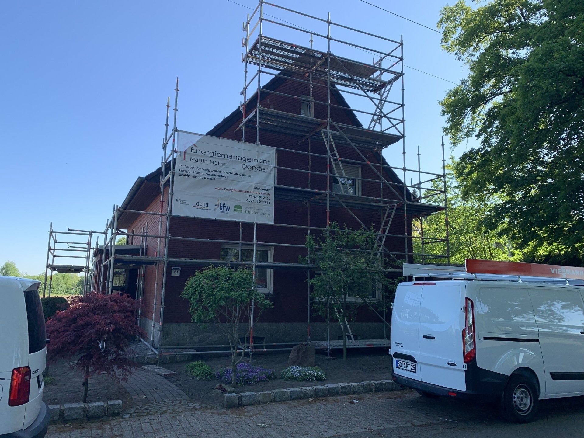 Rotes Haus im Bau, umgeben von Gerüsten. Zwei weiße Lieferwagen parken in der Nähe.