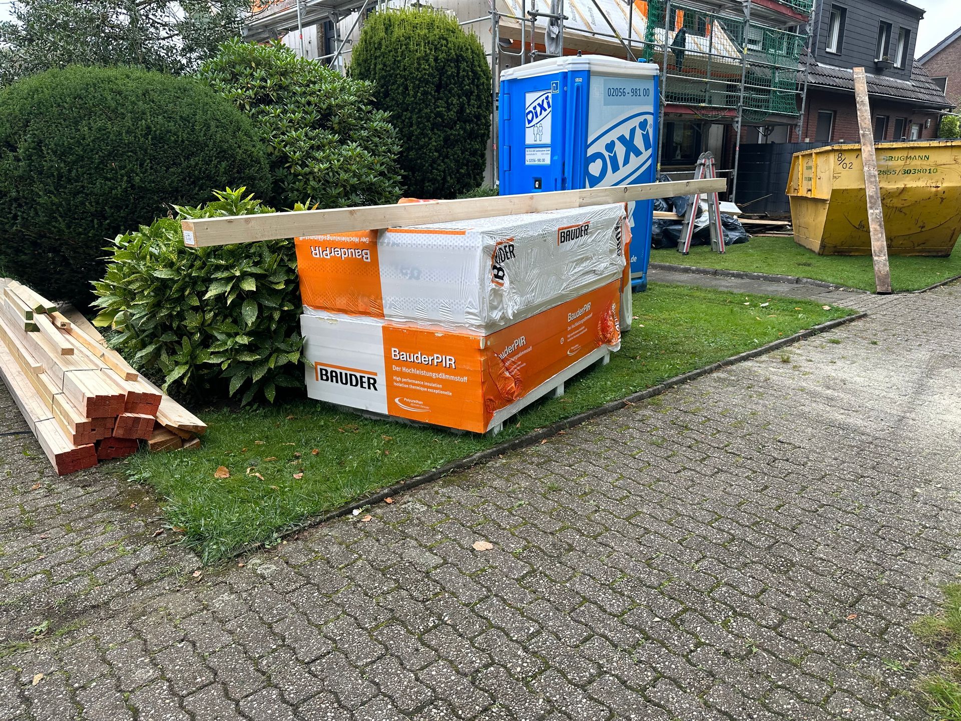 Baumaterialien auf Gras; orangefarbene Isolierung, Holzbalken, tragbare Toilette und ein Müllcontainer vor einem Haus.