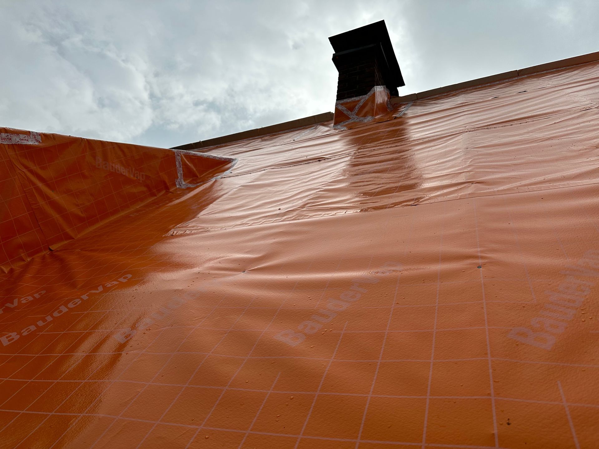Eine orangefarbene Plane bedeckt ein Dach mit einem schwarzen Schornstein vor einem bewölkten Himmel.
