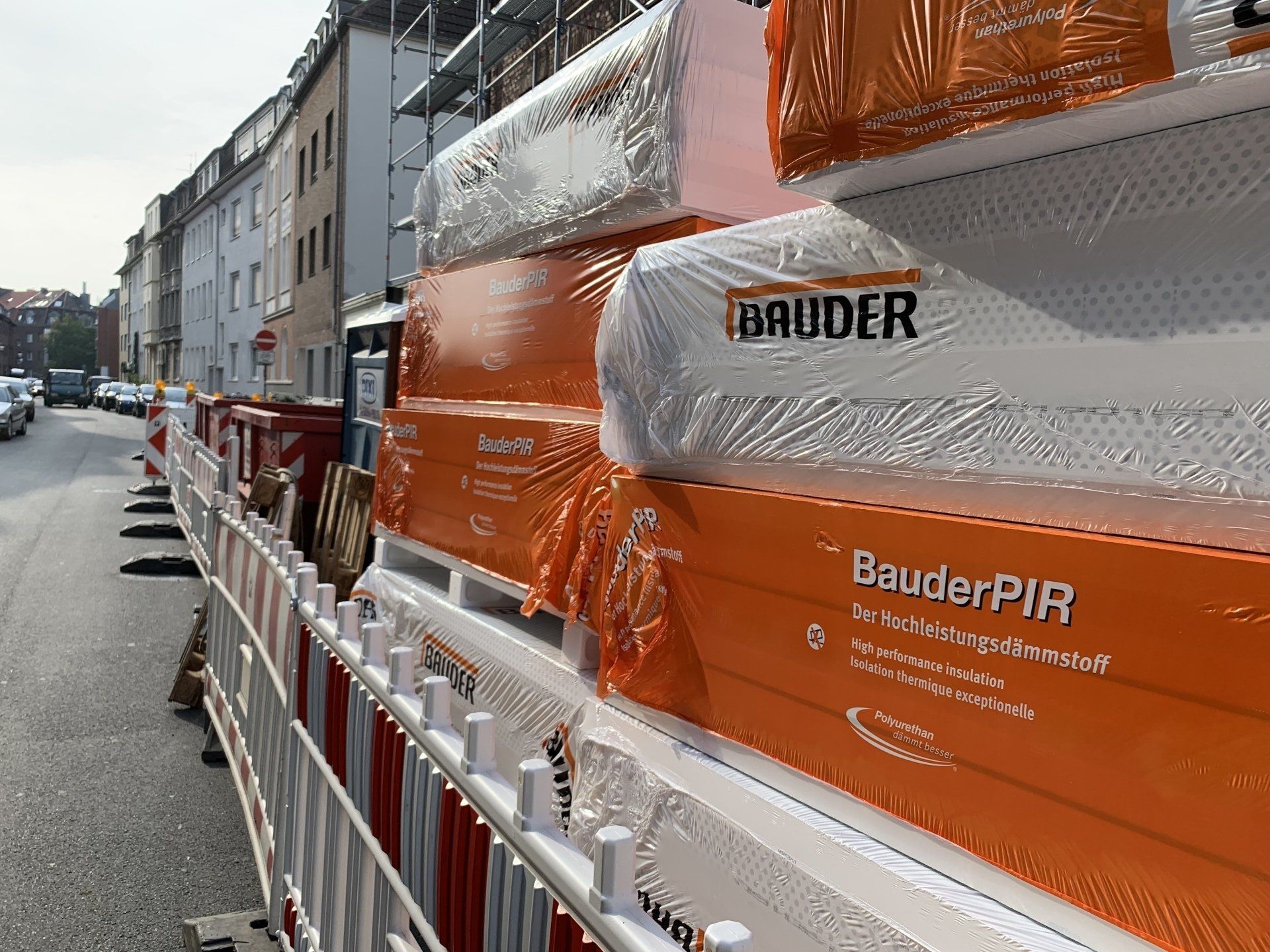 Orangefarbene und weiße Bauder-Isolierung, gestapelt hinter einer Baubarriere auf einer Stadtstraße.