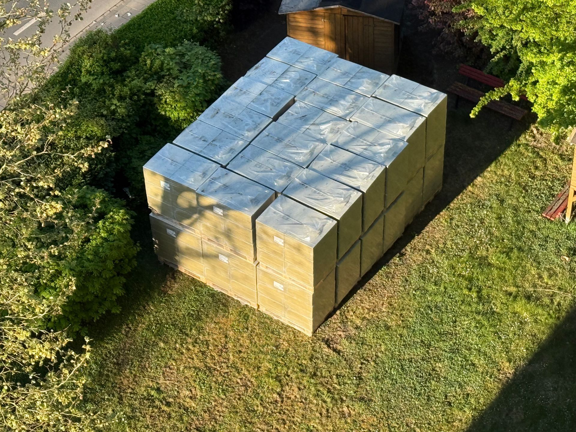 Ein Stapel rechteckiger Dämmblöcke in einem grasbewachsenen Hof neben Büschen und einem Schuppen.