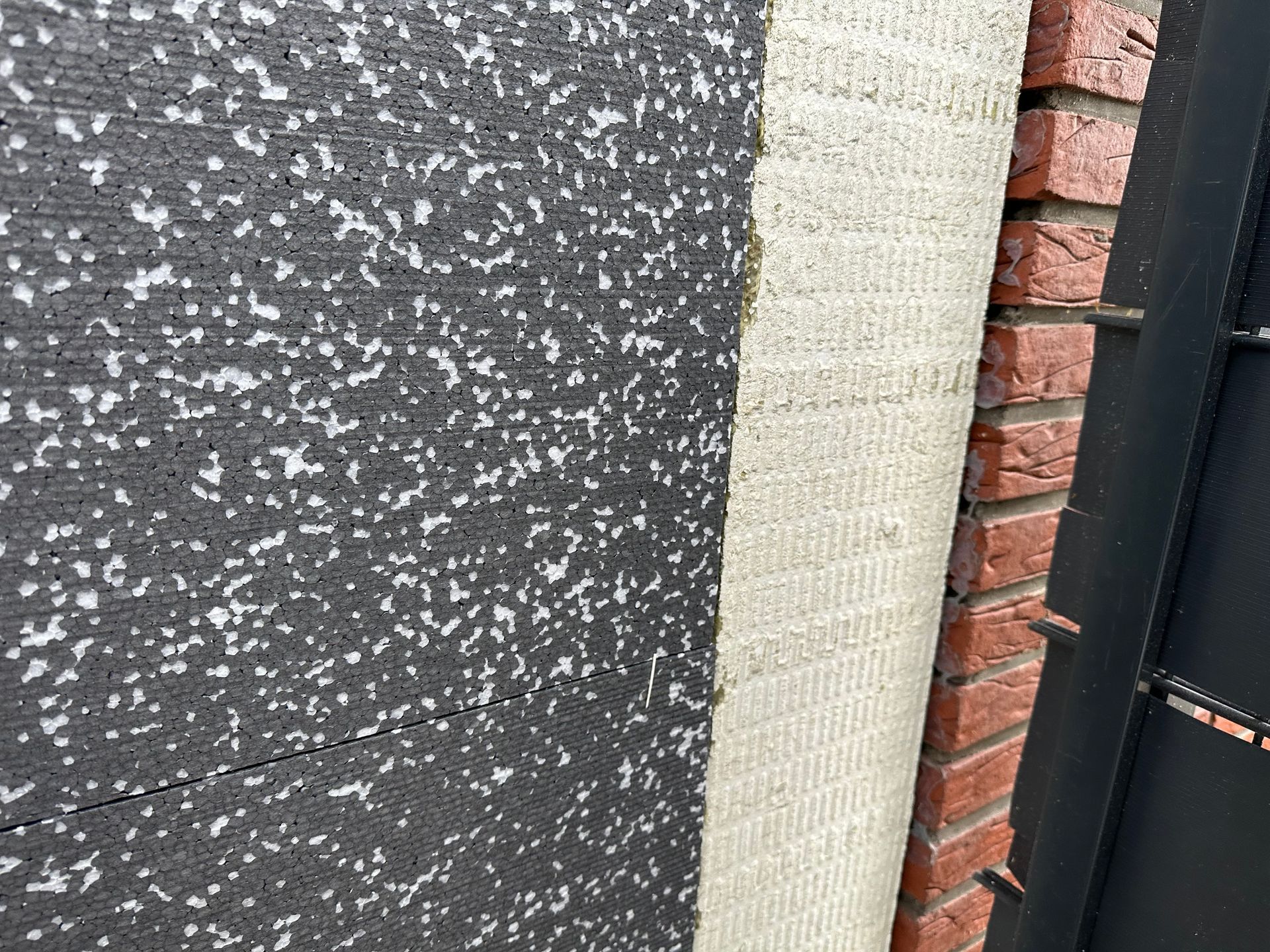 Nahaufnahme der Außenwand mit schwarzer Isolierung, strukturierter hellbeiger Beschichtung und rotem Backstein.