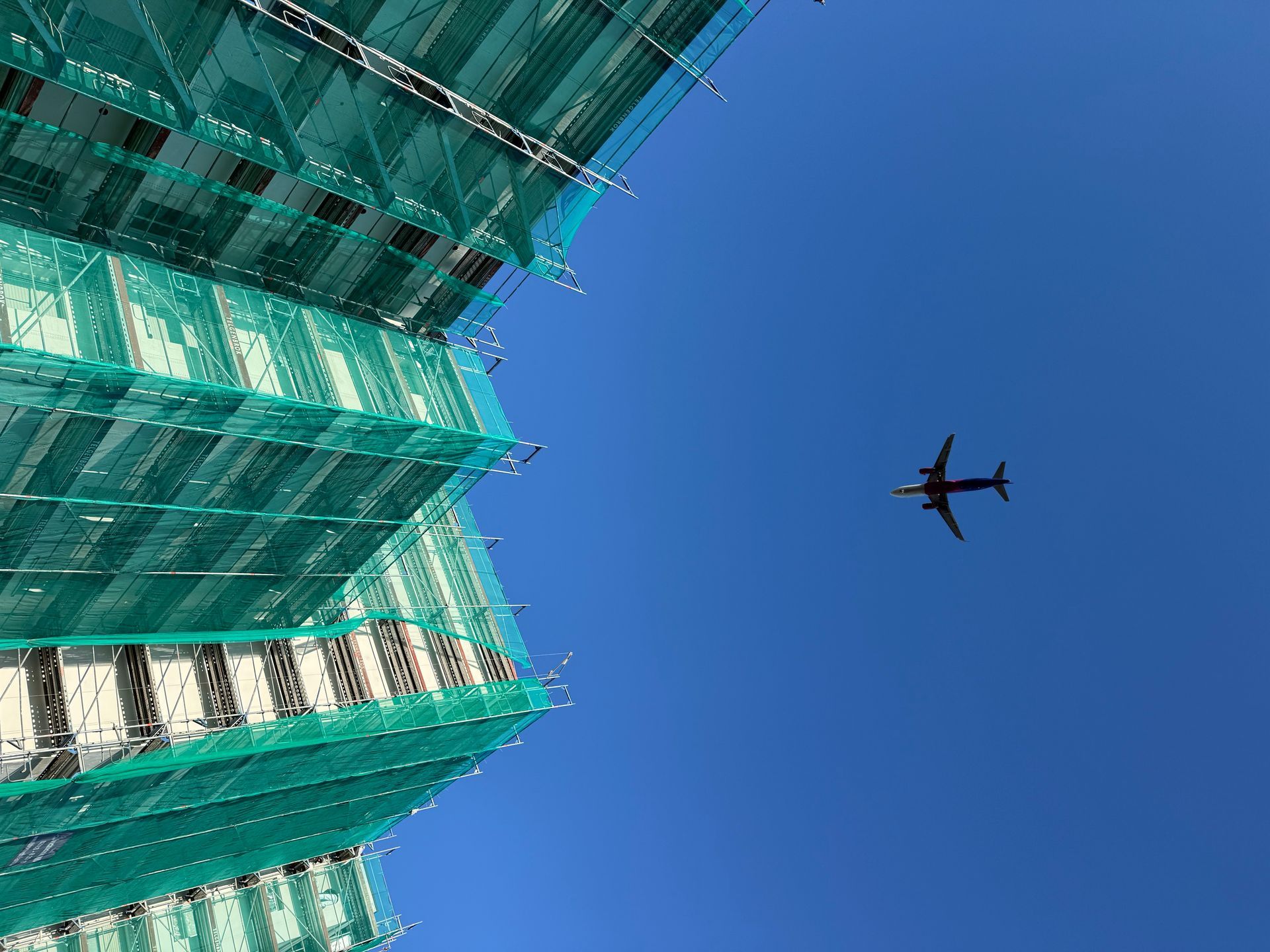 Gebäude im Bau mit grünem Sicherheitsnetz vor einem klaren blauen Himmel; Flugzeug im Flug.