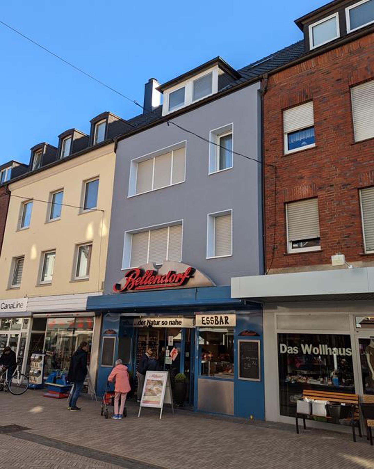 Schaufenster mit rotem Schild „Restaurant“, hellblaue Fassade, Fußgänger und Geschäfte an einem sonnigen Tag.