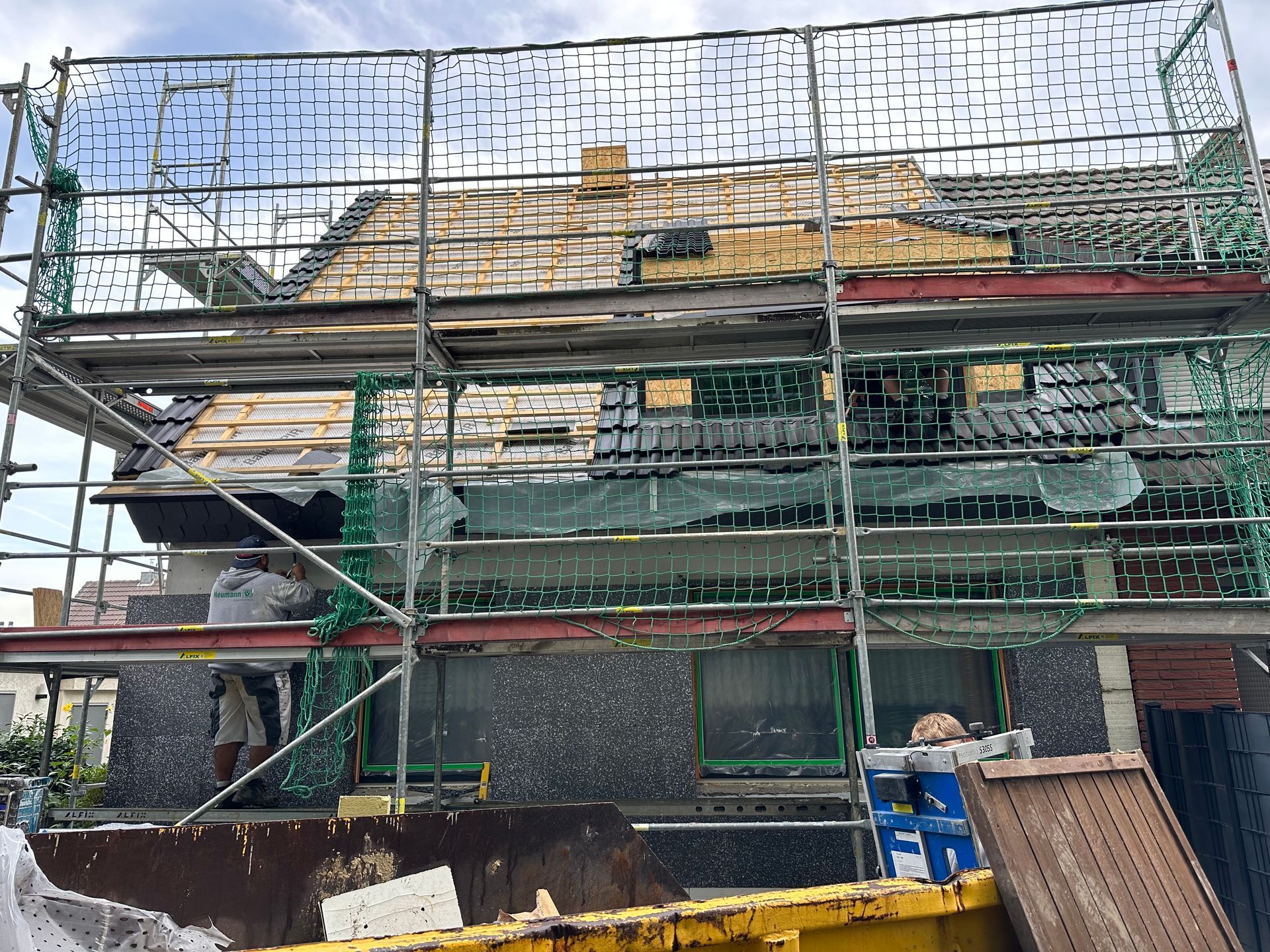 Haus im Bau, Dach teilweise entfernt, Gerüst, Sicherheitsnetz, Mülltonnen.