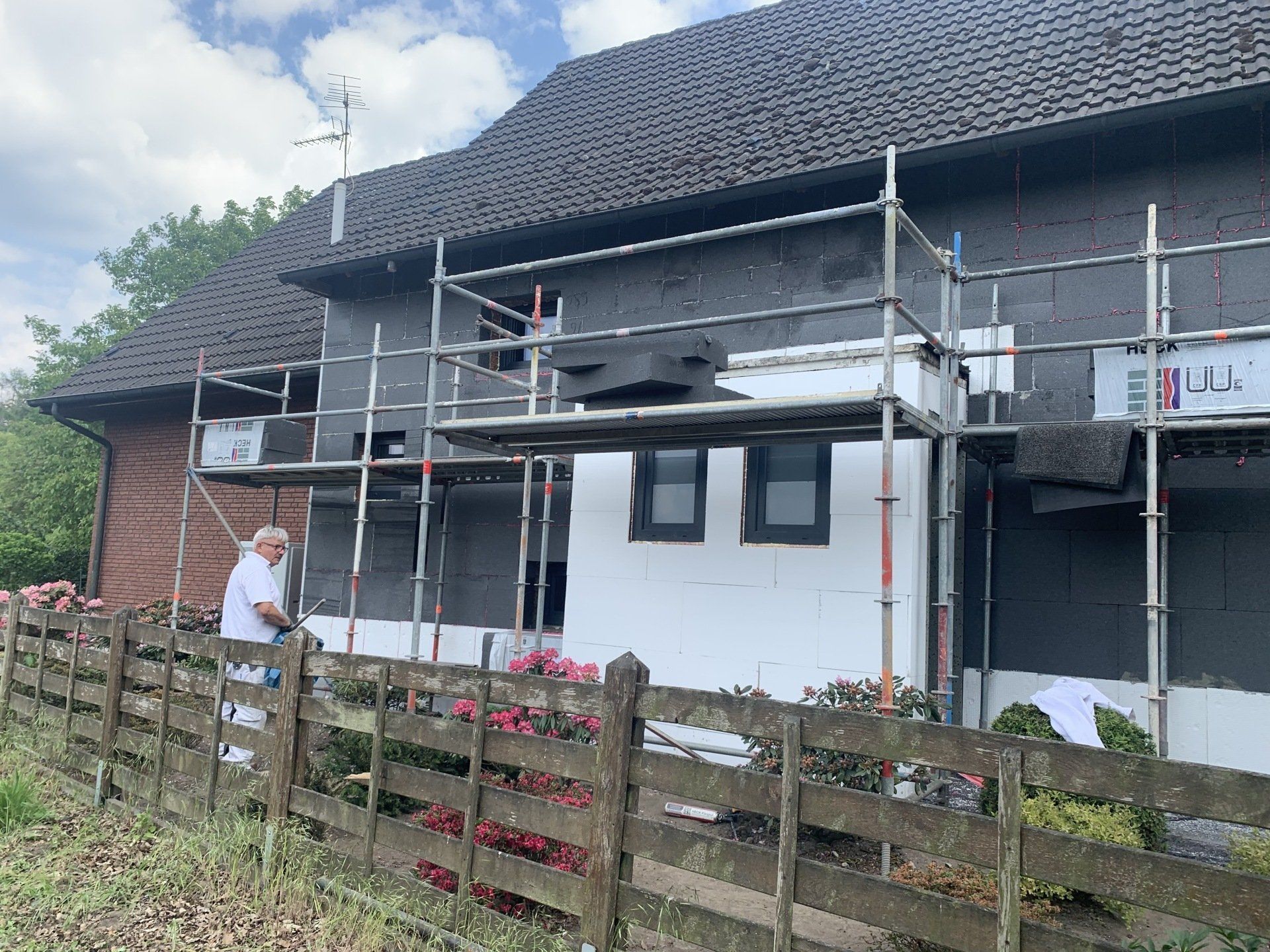 Mann arbeitet mit Gerüst an der Hausfassade; weiße und schwarze Paneele, Holzzaun und Rosen im Garten.