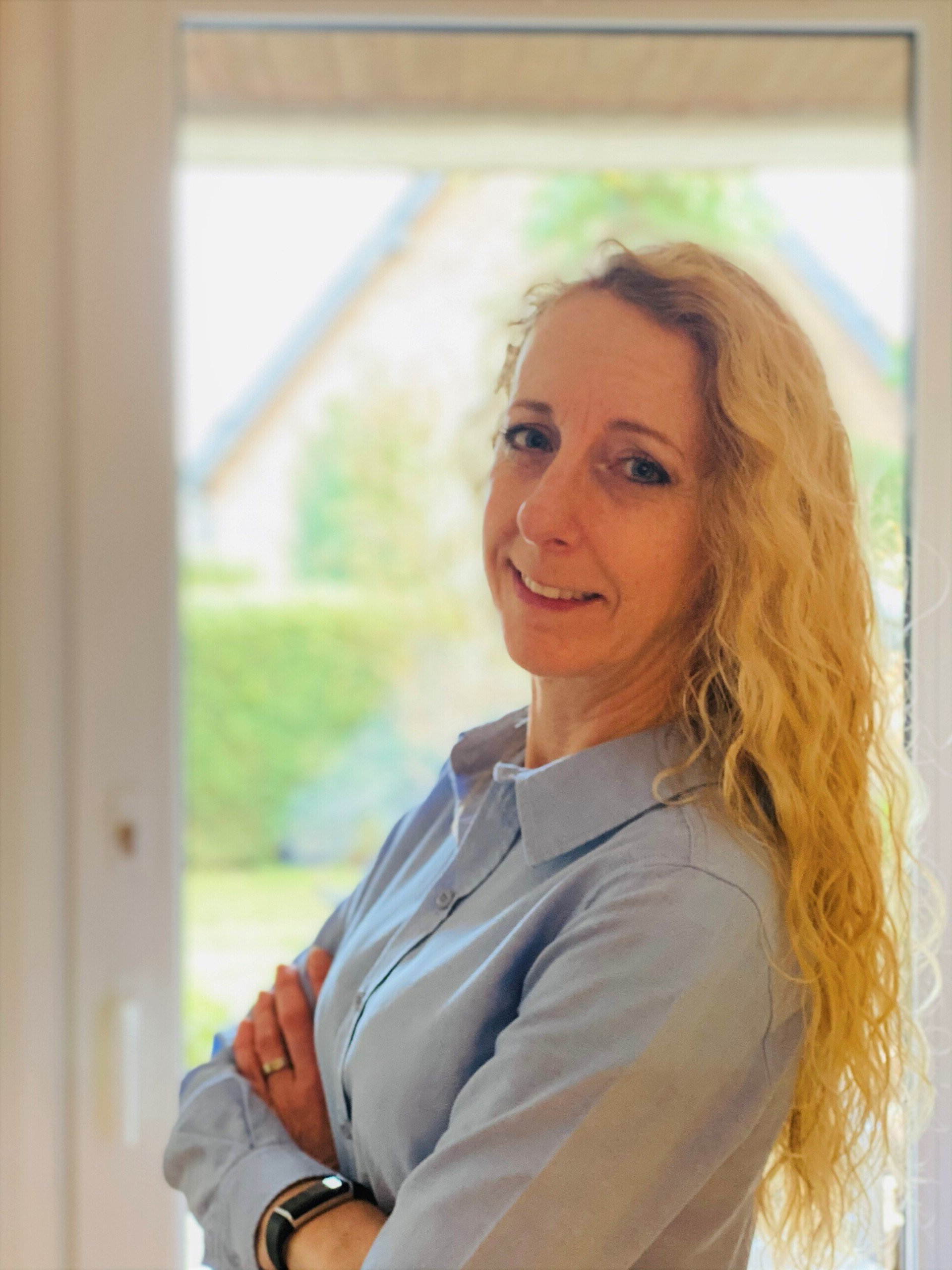 Frau mit blonden Locken, verschränkten Armen, lächelt vor einem Fenster, zartblaues Hemd.
