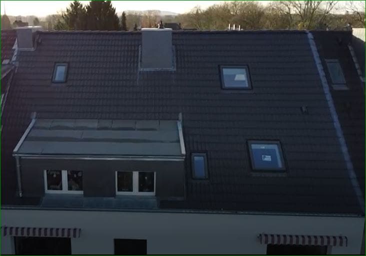 Dunkelgraues Dach mit Schornsteinen und Oberlichtern; Flachdachabschnitt links.