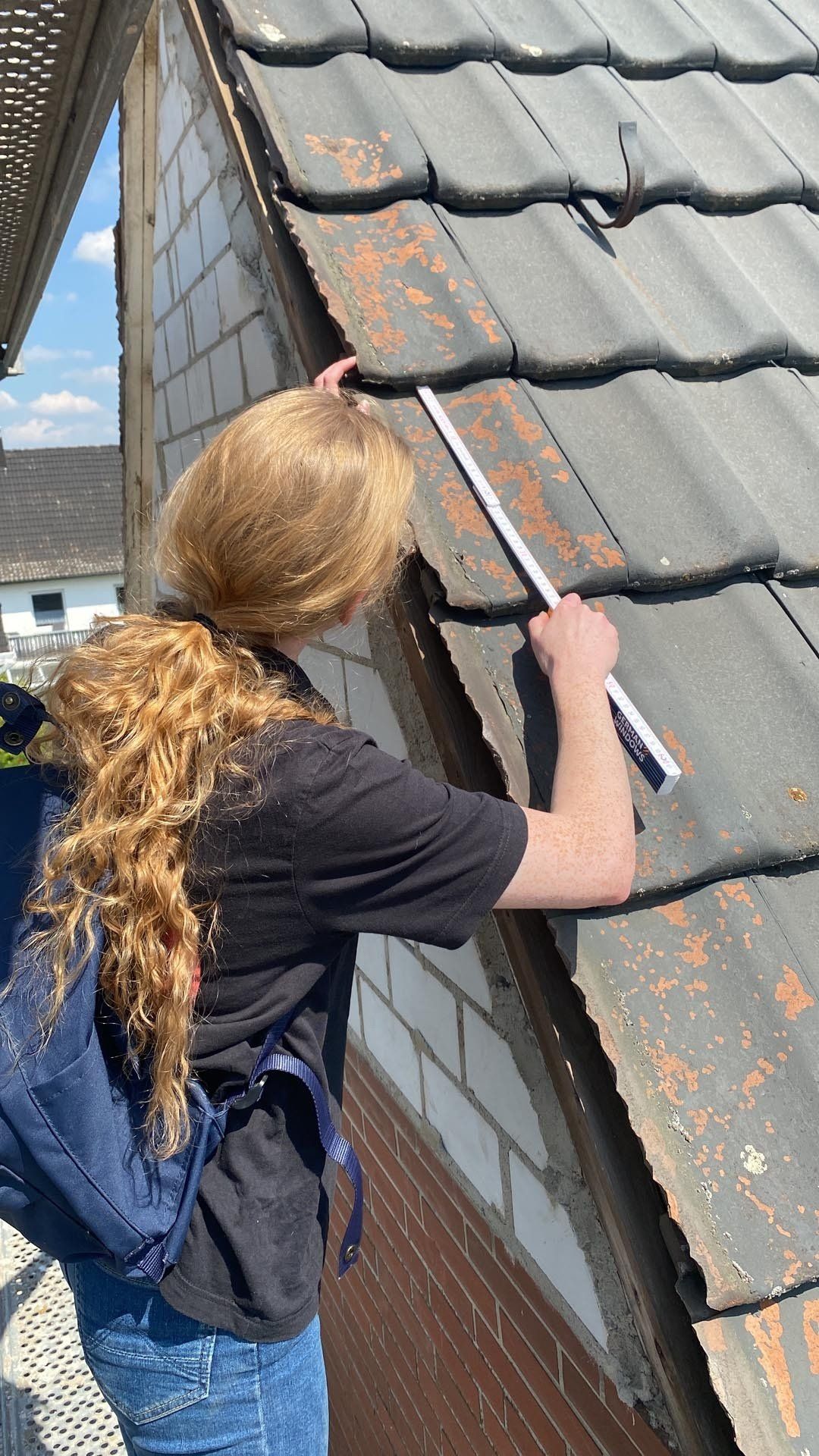 Person mit langem, lockigem Haar, die an einem sonnigen Tag im Freien mit einem Lineal die Kante eines Daches misst.