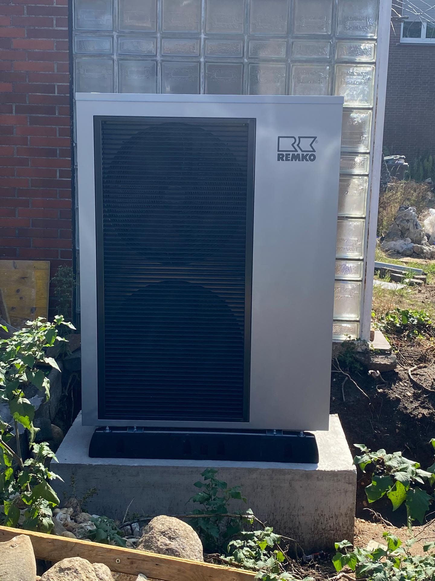 Eine silberne Wärmepumpe in einem Garten, montiert auf einem Betonsockel vor einer Glasbausteinwand.