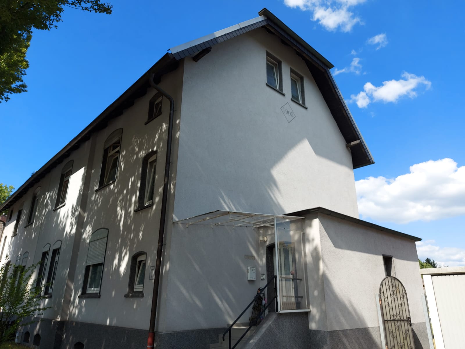 Zweistöckiges graues Gebäude mit dunklem Dach vor blauem Himmel mit Wolken.