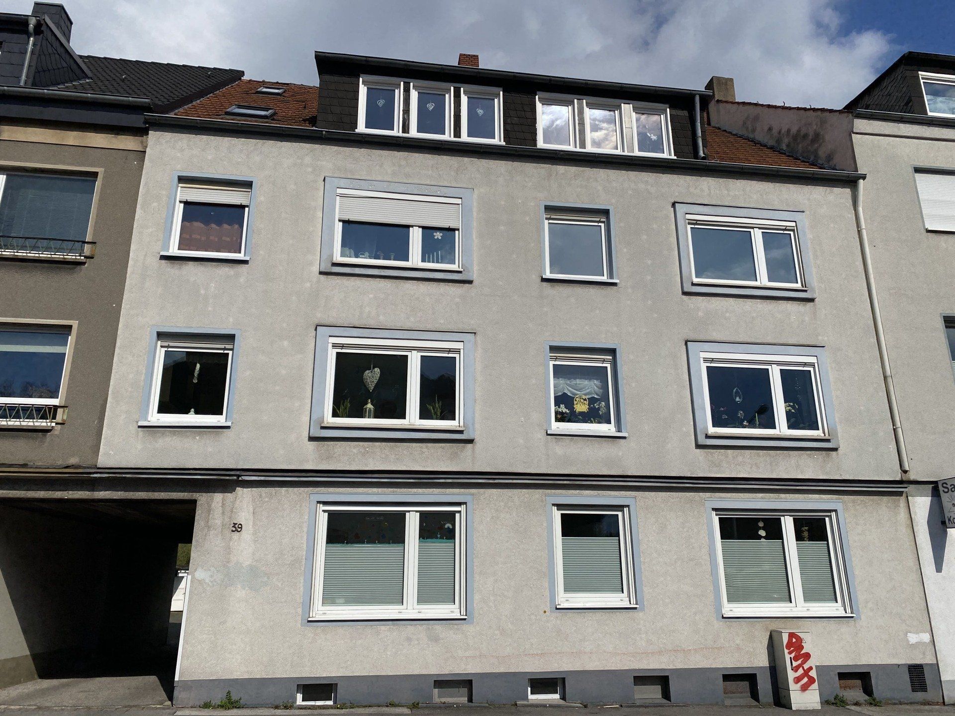 Graues Mehrfamilienhaus mit mehreren weiß gerahmten Fenstern, dunklem Dach und überdachtem Eingang.