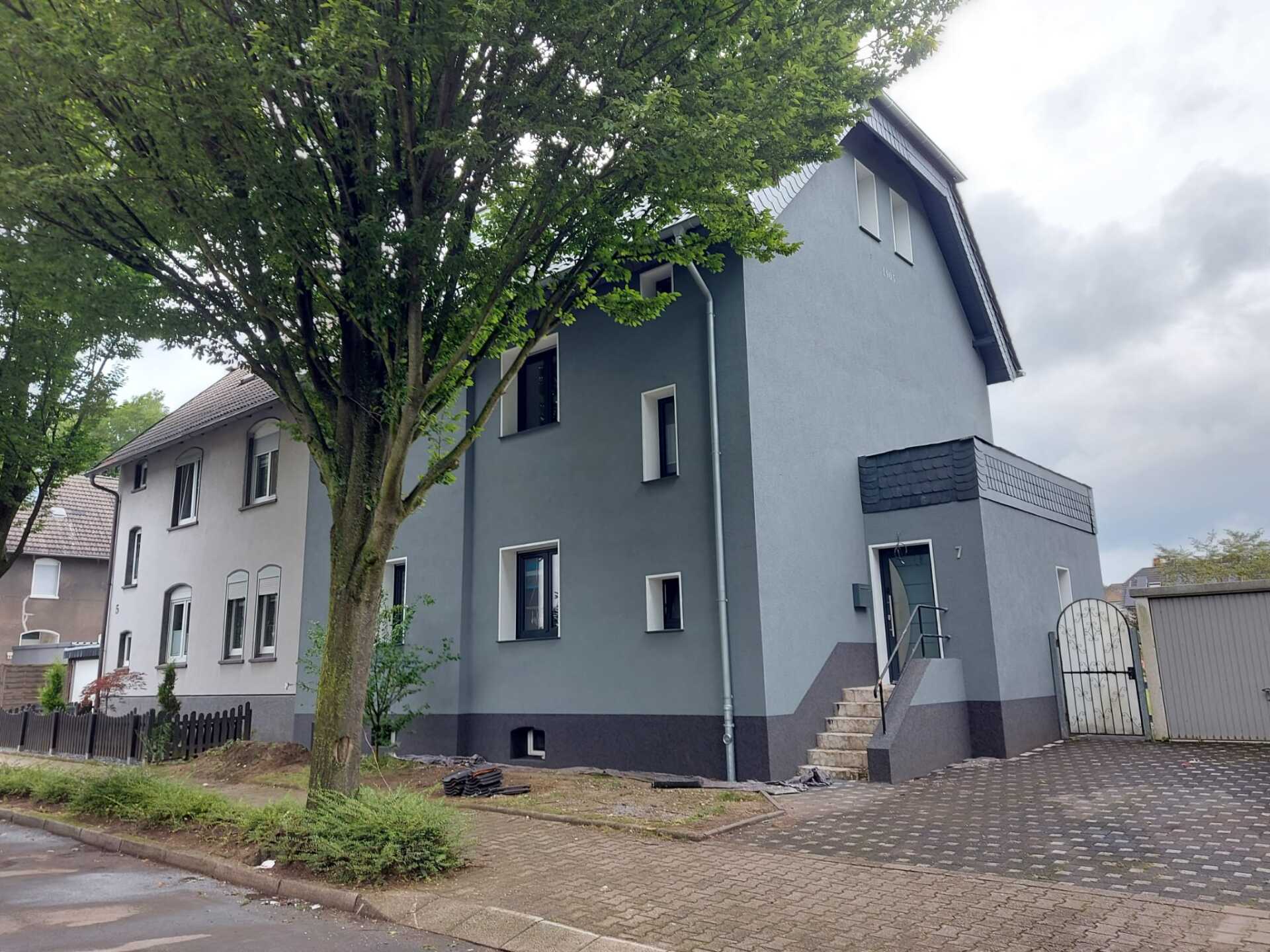 Graues Haus mit dunkelgrauen Verzierungen und einem Baum davor.