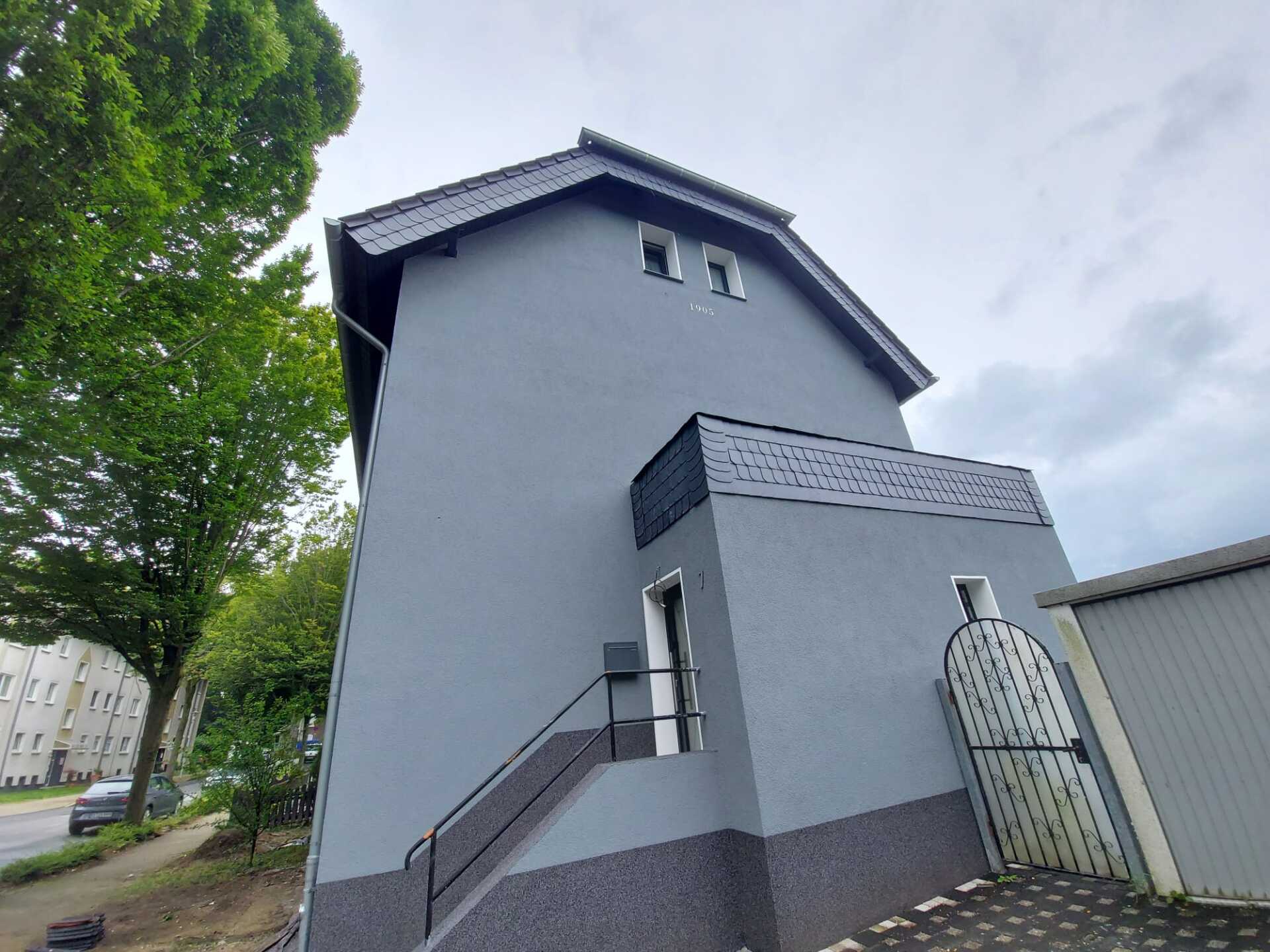 Grau gestrichenes Gebäude mit dunklem Dach, kleinem Balkon und einer Treppe, die zu einer Tür führt.