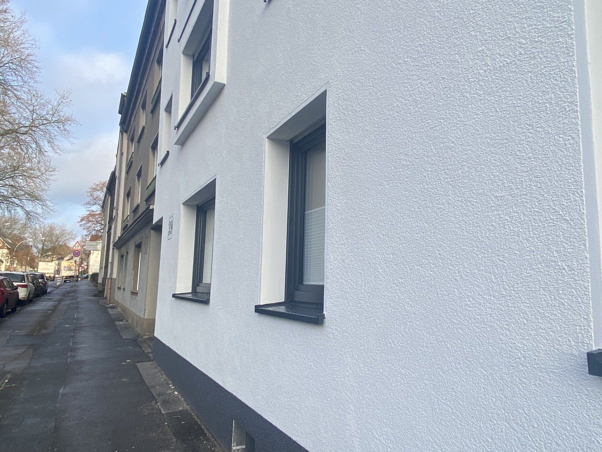Außenansicht eines weißen Stuckgebäudes mit dunklen Fensterrahmen, neben einer Straße mit geparkten Autos.