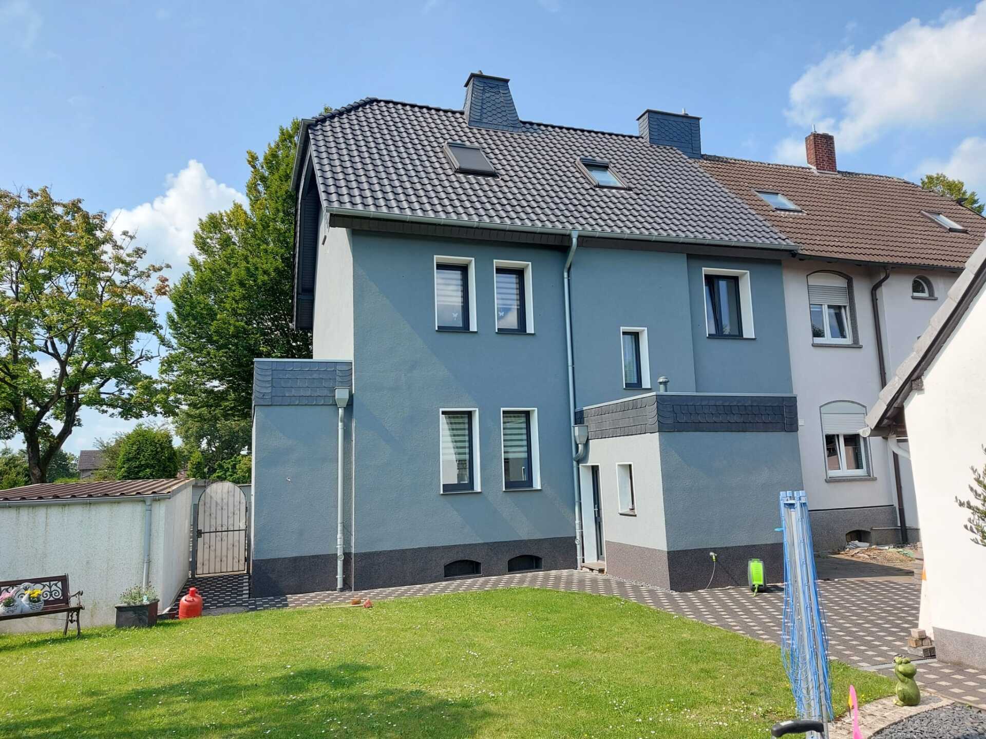 Rückansicht eines blauen zweistöckigen Hauses mit schwarzem Dach und kleinem Garten an einem sonnigen Tag.