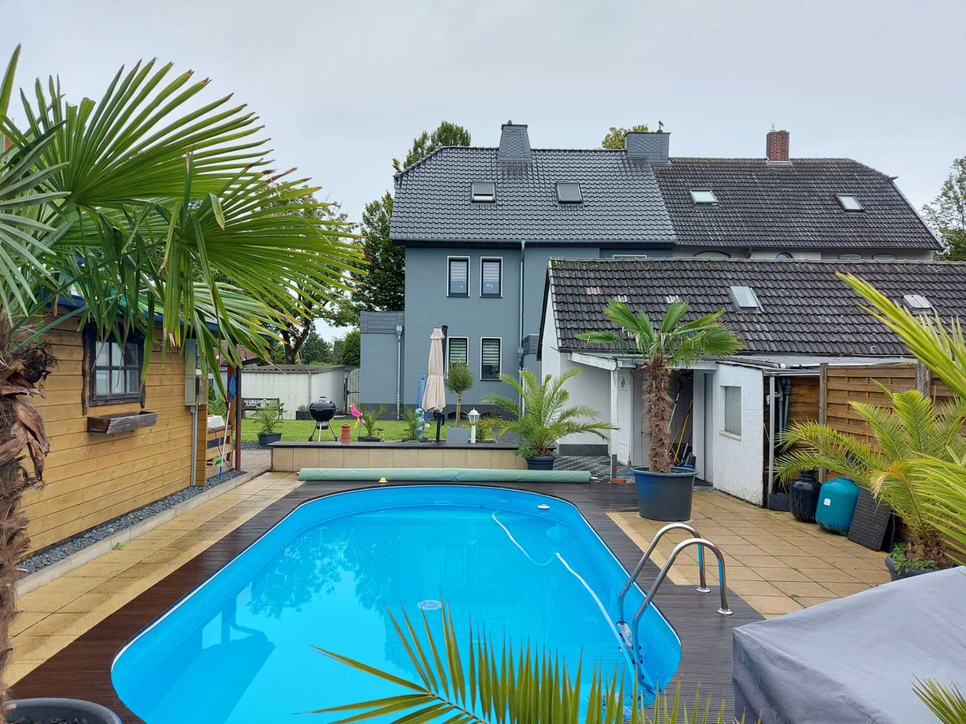 Hinterhof mit Pool, Palmen und zweistöckigem grauen Haus unter einem bewölkten Himmel.
