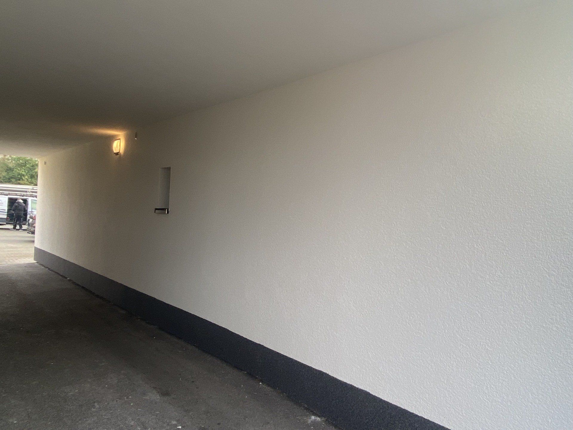 Tunnel mit weißen strukturierten Wänden, schwarzer Fußleiste und einer Leuchte. Asphaltboden.