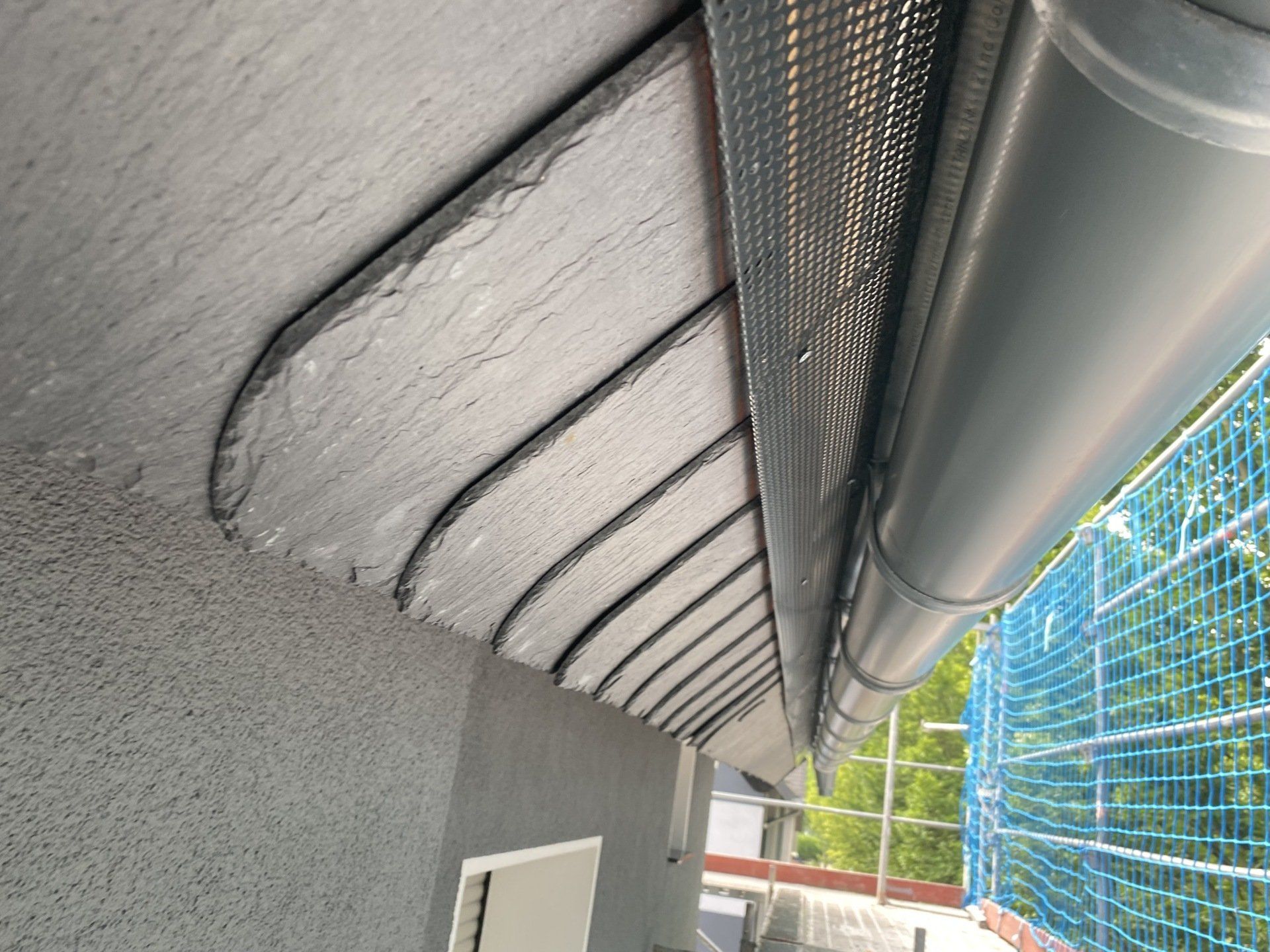 Schieferdachkante mit Dachrinne und Sichtschutz, vor grauer Stuckwand, von unten gesehen.