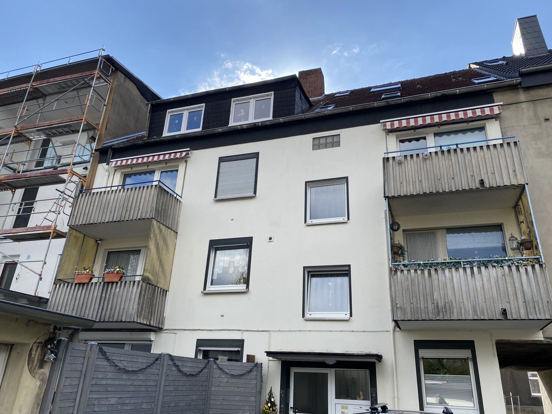 Außenansicht eines Mehrfamilienhauses mit Balkonen, weißen Wänden und dunklem Dach vor blauem Himmel.