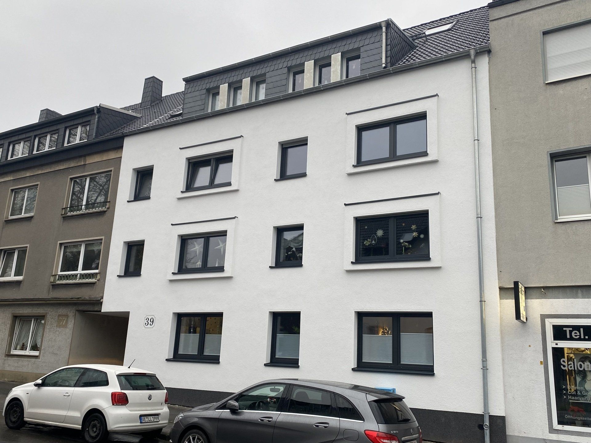 Weißes Wohnhaus mit schwarz gerahmten Fenstern und davor geparkten Autos. Grauer, bewölkter Himmel.