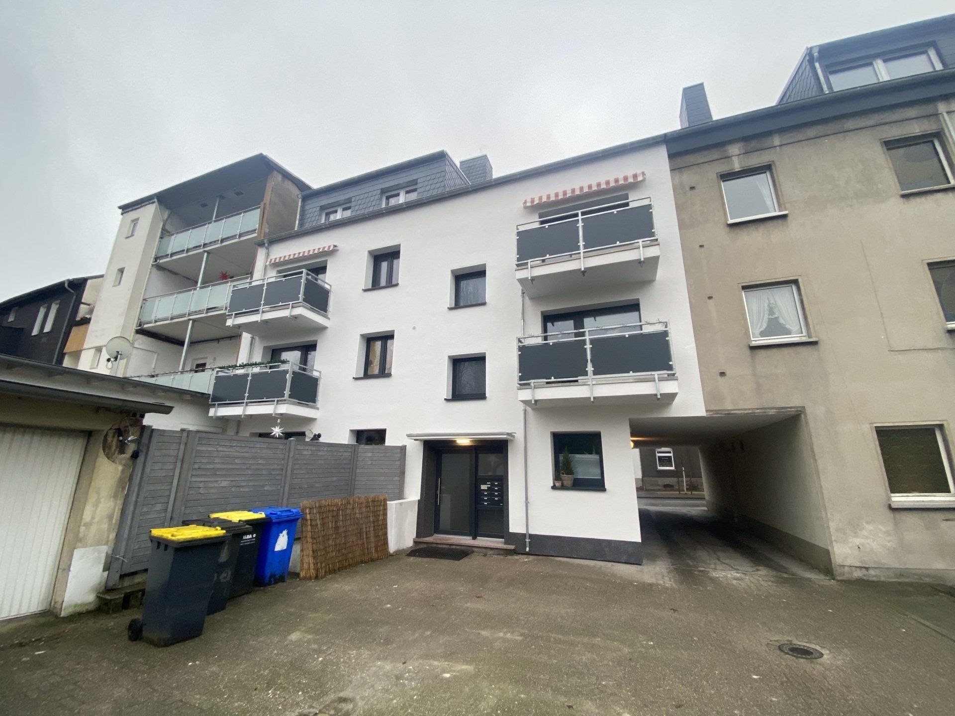 Mehrfamilienhaus mit weißer Fassade und grauen Balkonen, Eingang und Mülltonnen. Bewölkter Himmel.