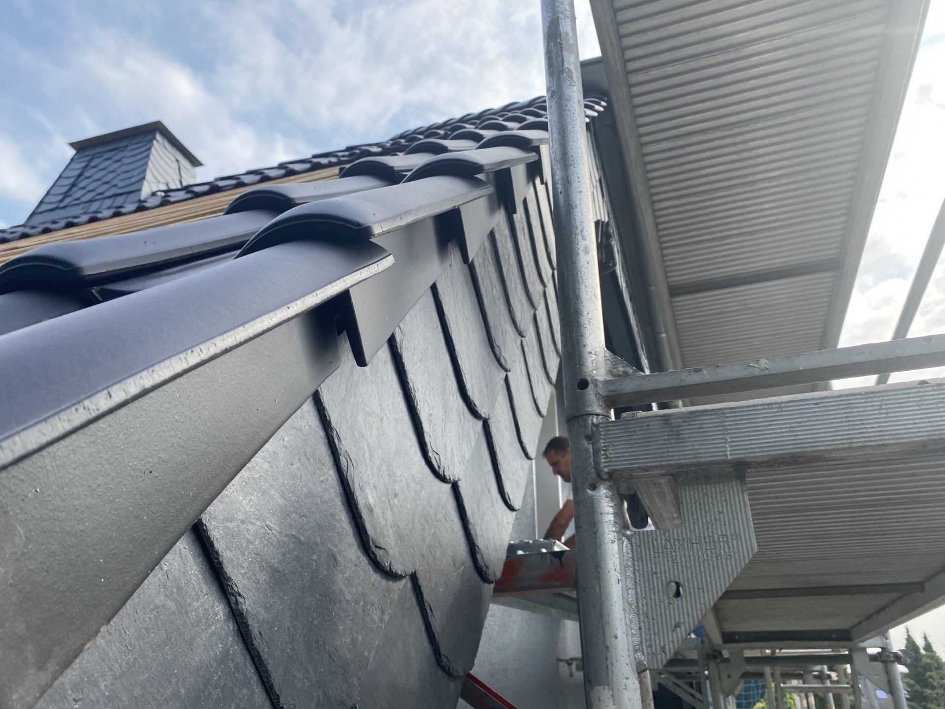 Dachdecker auf Gerüst repariert Schieferdach; Himmel und Schornstein sichtbar.