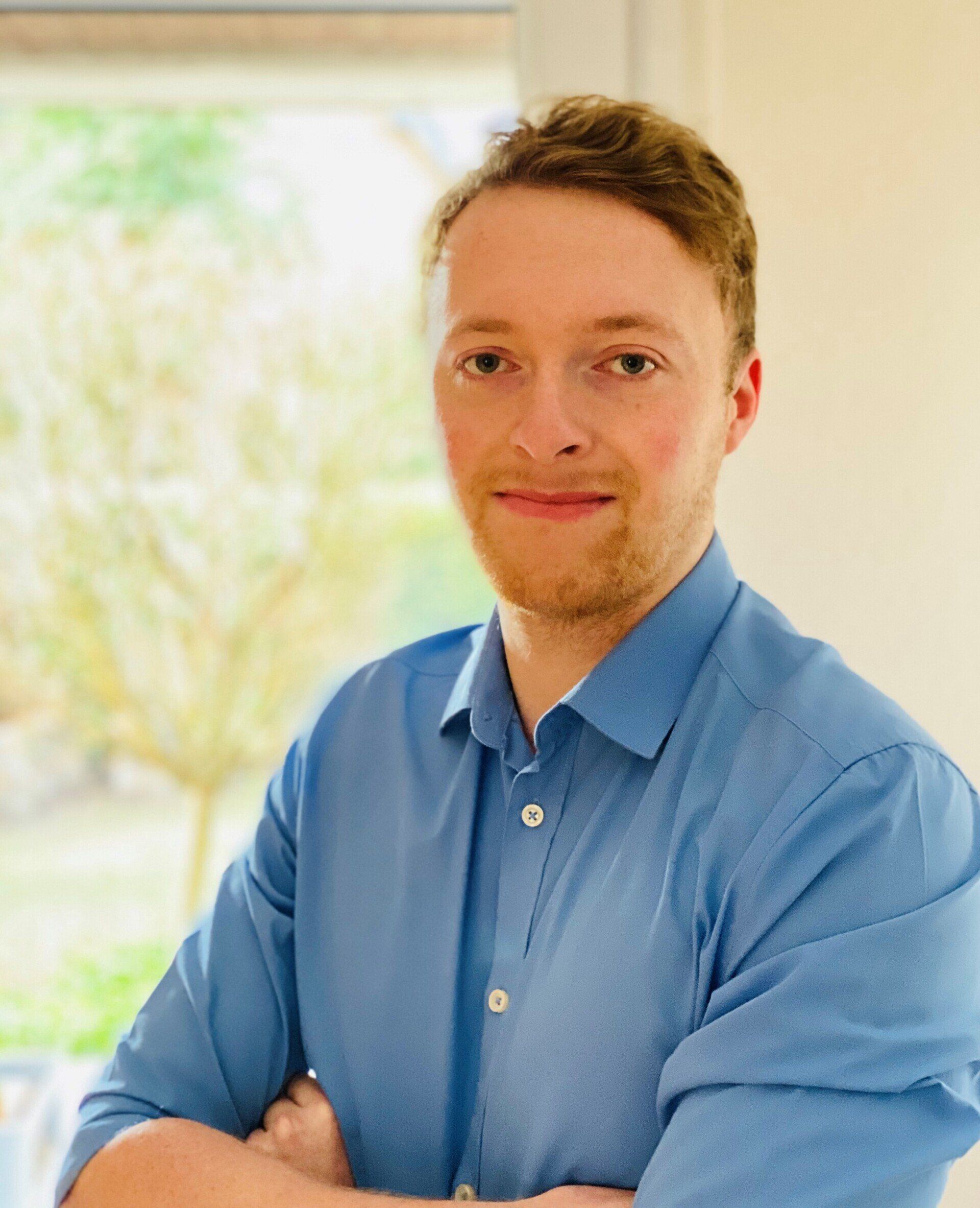 Mann mit blondem Haar und Bart, trägt ein blaues Button-Down-Hemd, die Arme verschränkt, lächelnd.