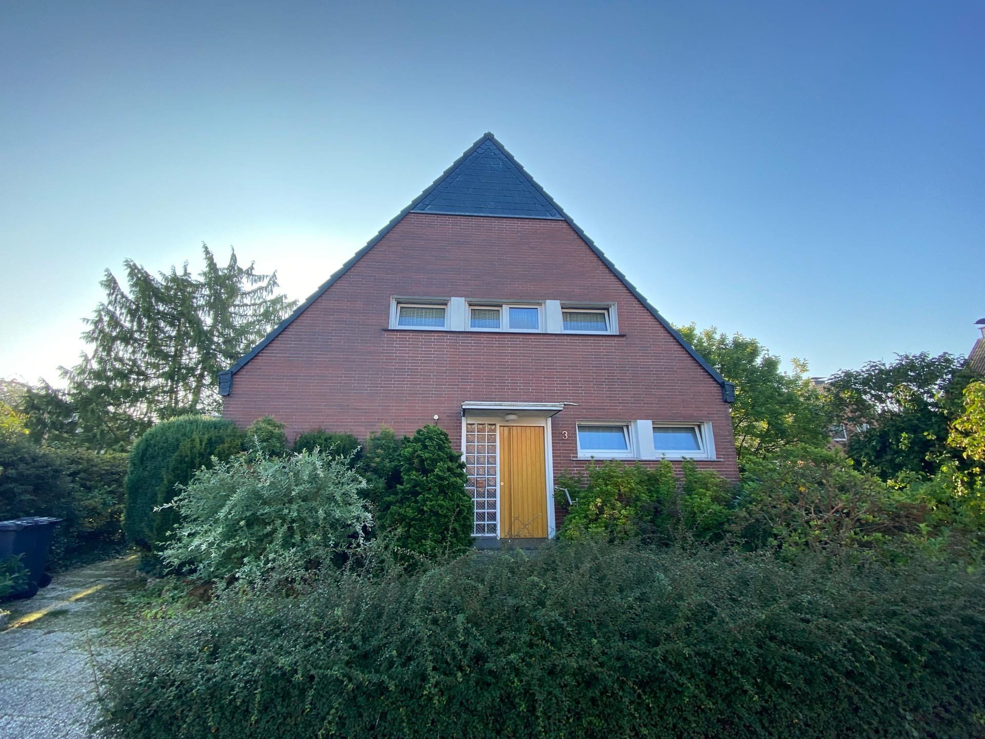 Ein pyramidenförmiges Backsteinhaus mit dunklem Dach und gelber Tür, umgeben von Büschen.