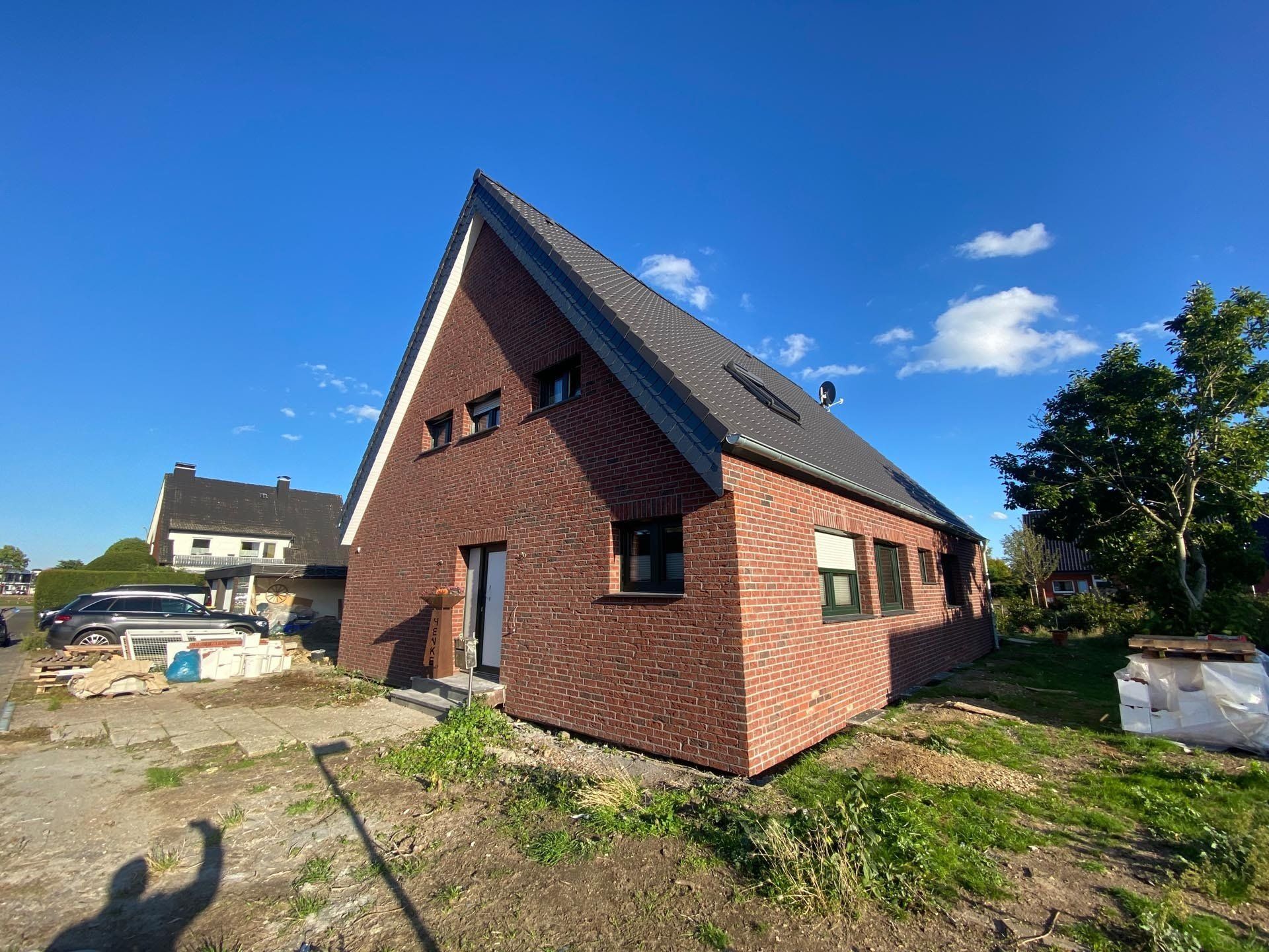 Backsteinhaus mit schrägem Dach unter blauem Himmel. Baustelle, kleine Fenster und ein Baum.