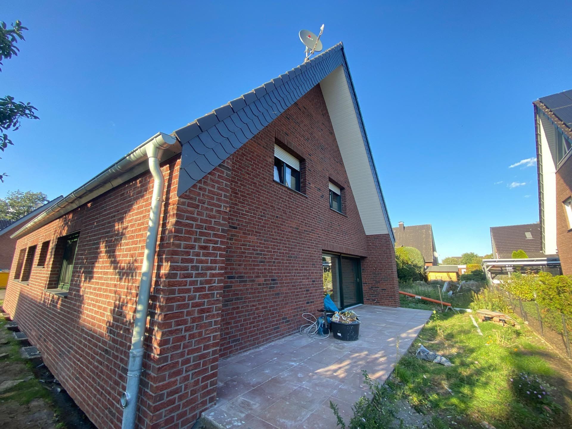 Backsteinhaus mit dunklem Dach, weißen Zierleisten und einer Betonterrasse an einem sonnigen Tag.