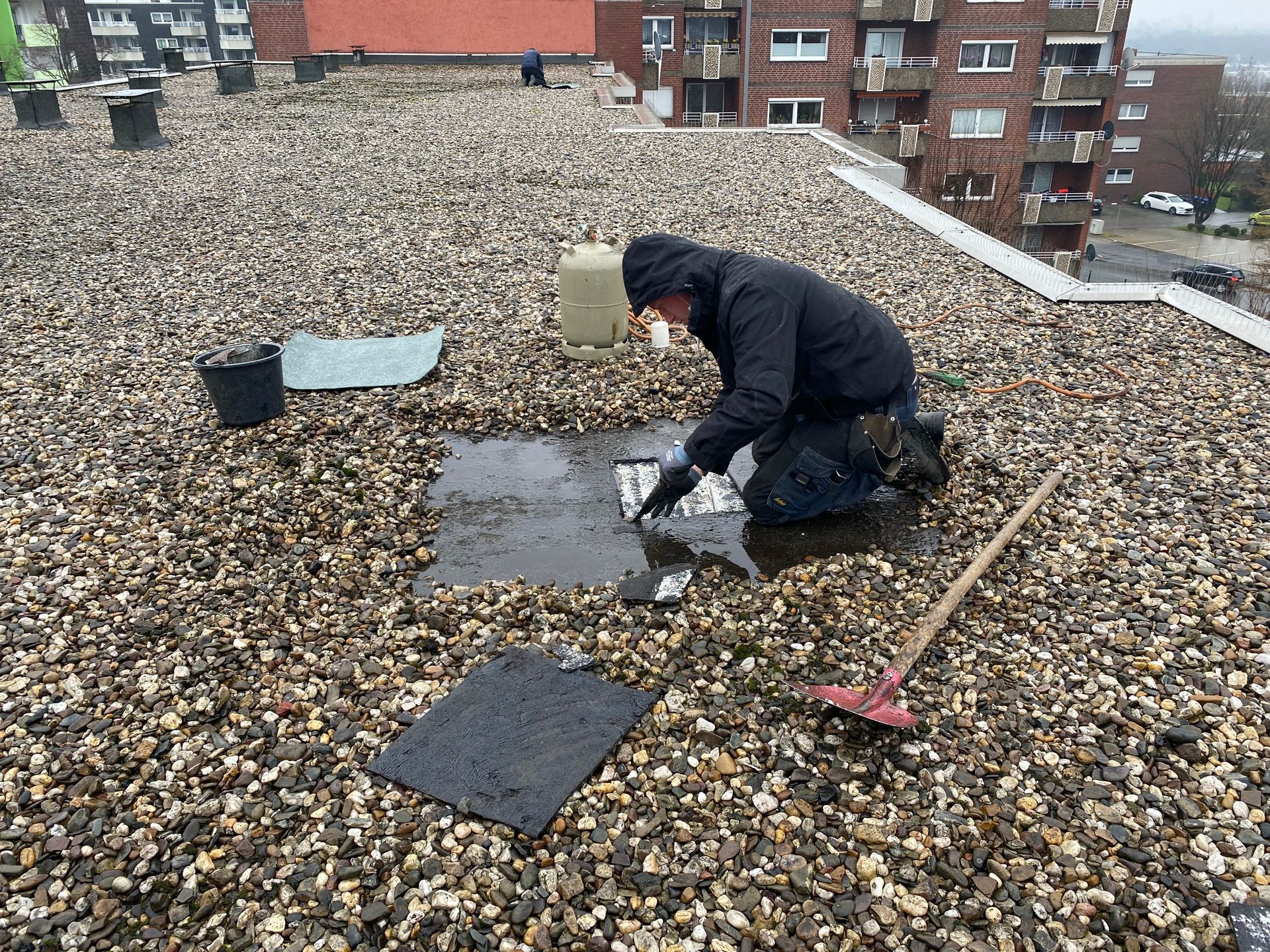 Person auf einem Kiesdach, die ein Leck repariert, Gebäude im Hintergrund.