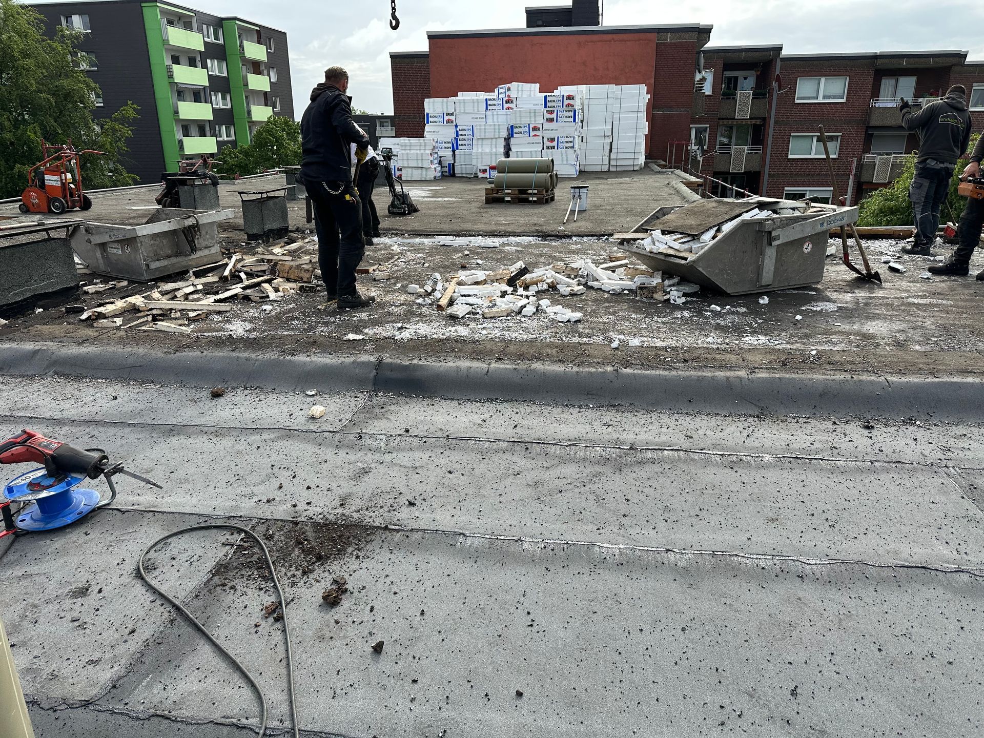 Arbeiter entfernen Schutt auf einem Dach. Im Hintergrund sind ein grünes Wohnhaus und ein rotes Backsteingebäude zu sehen.