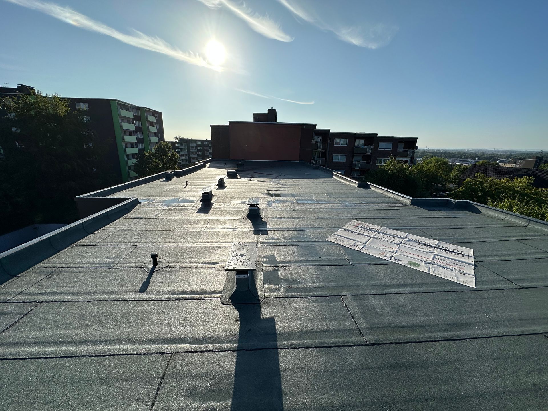 Blick auf ein Flachdach an einem sonnigen Tag. In der Ferne sind Gebäude und Bäume unter einem blauen Himmel sichtbar.