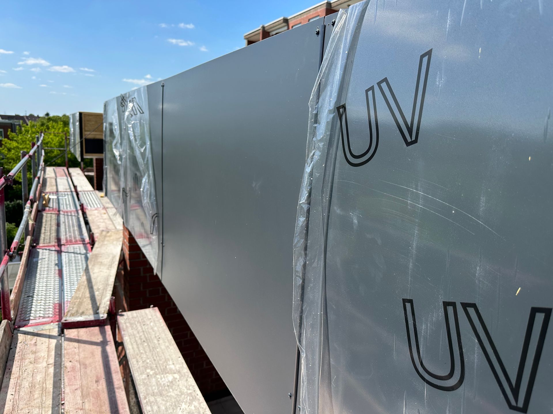 Baustelle mit grauen, durch UV-Folie geschützten Metallplatten an einer Ziegelwand. Links ein Gerüst.