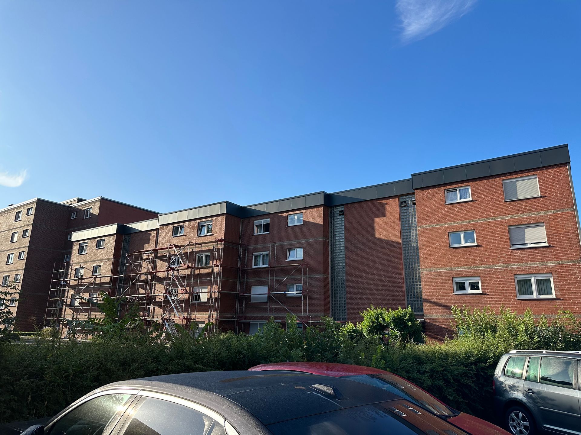 Backsteinwohnhaus mit Gerüst unter blauem Himmel. Autos und Büsche davor.