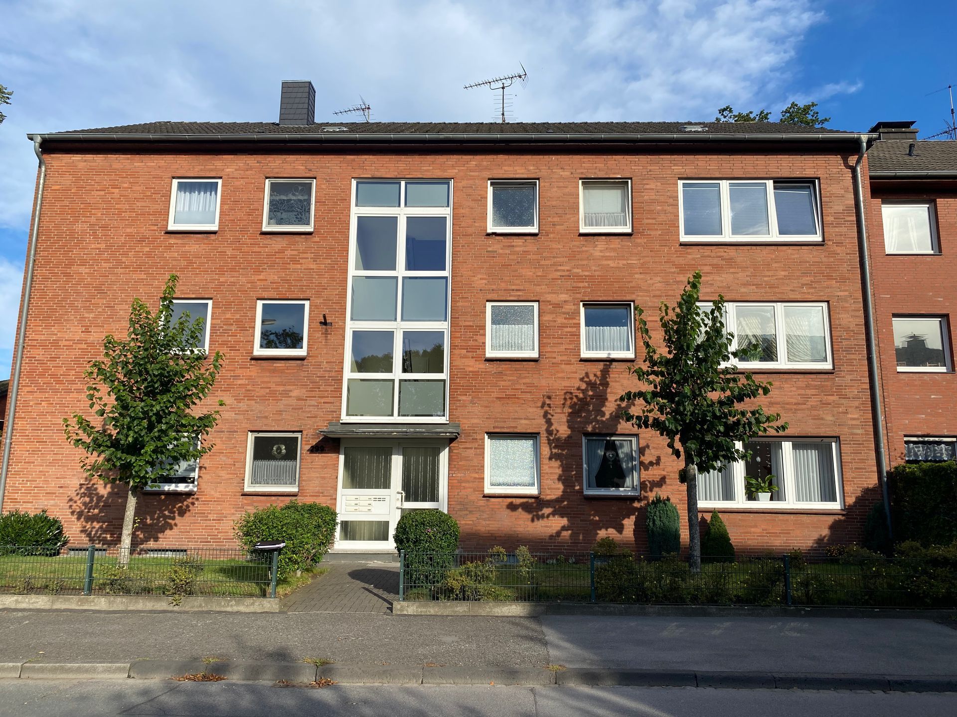 Backstein-Mehrfamilienhaus mit weißen Fensterrahmen und einem Glaseingang; davor Bäume.