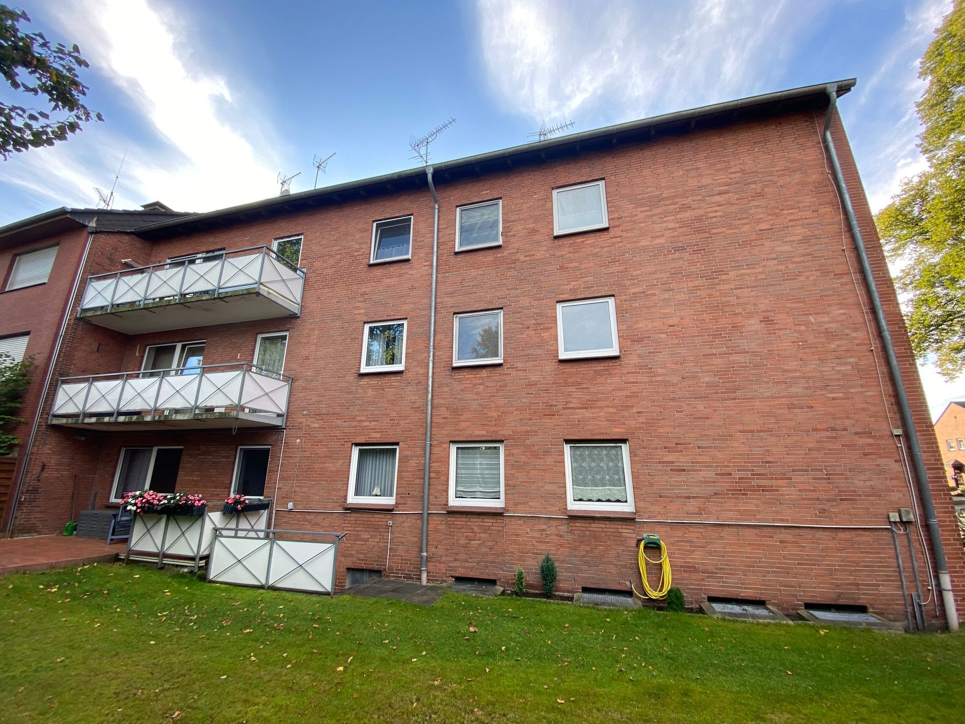 Dreistöckiges Backsteinwohnhaus mit Balkonen und Fenstern vor blauem Himmel mit etwas Grün im Vordergrund.