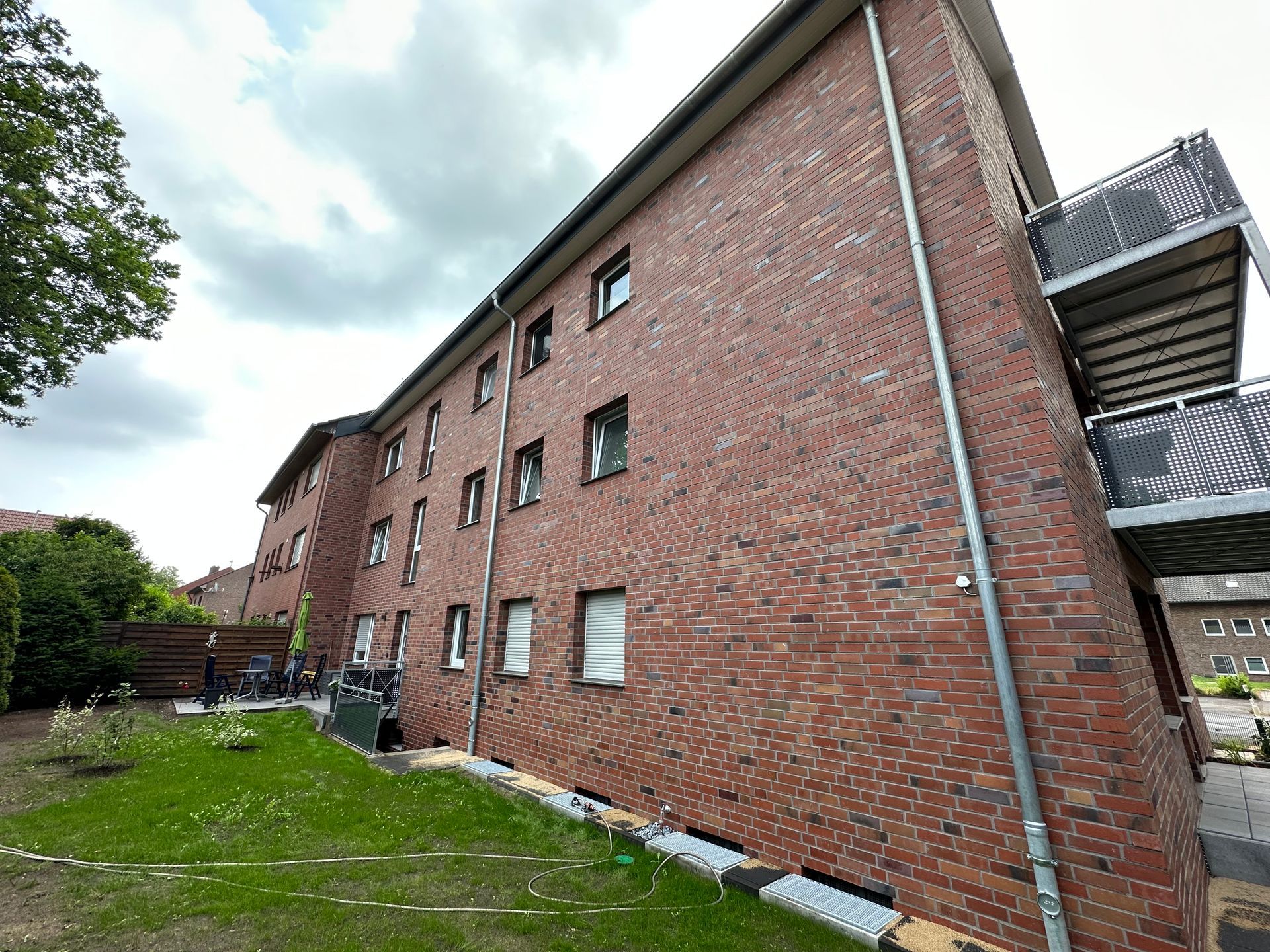 Wohnhaus aus rotem Backstein mit mehreren Fenstern, Außenansicht. Grünes Gras im Vordergrund, bewölkter Himmel.