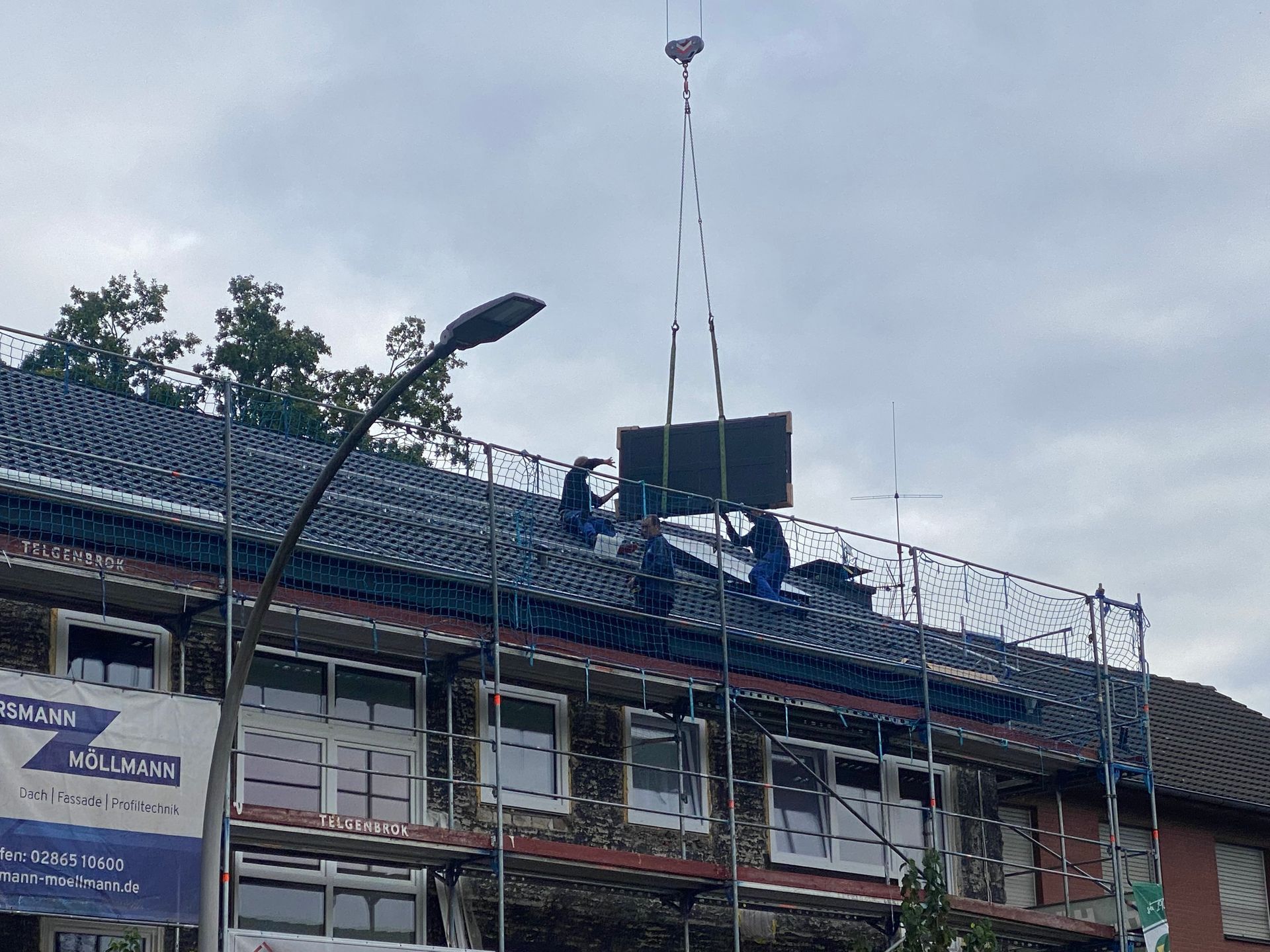Arbeiter installieren Solarmodule auf dem Dach unter bewölktem Himmel. Gerüst und Kran sind vorhanden.