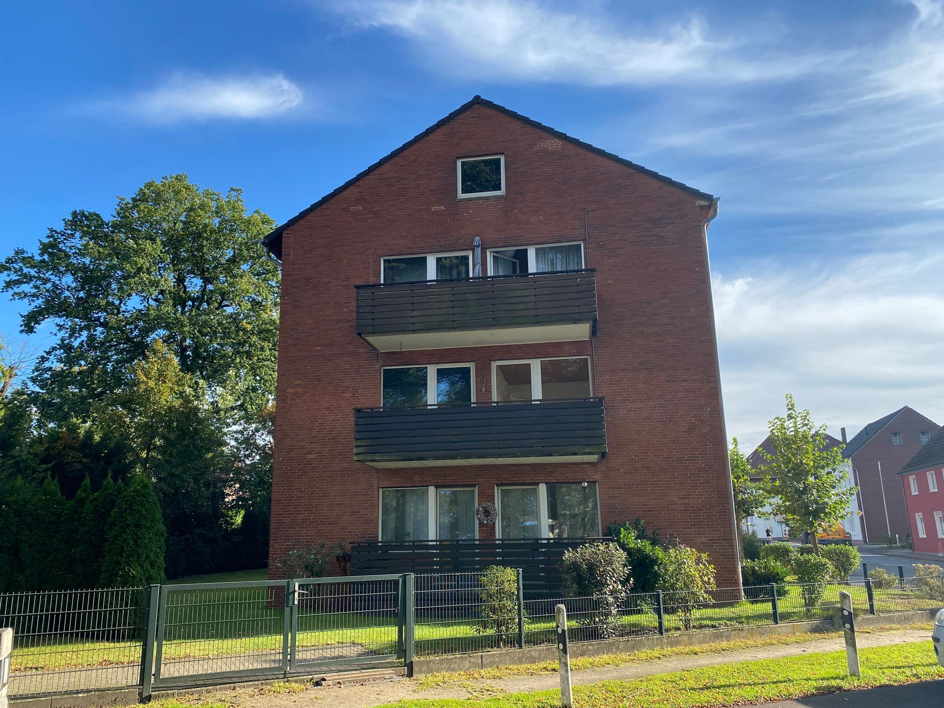 Wohnhaus aus rotem Backstein mit drei Balkonen, blauem Himmel und grünen Bäumen.