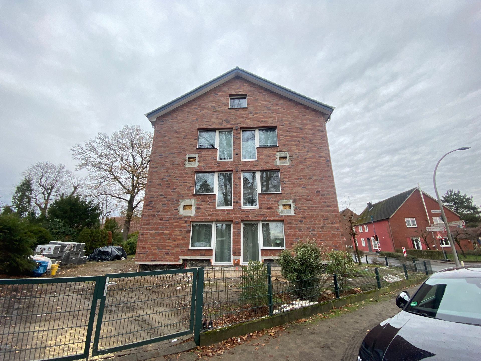 Wohnhaus aus rotem Backstein mit mehreren Fenstern, einem bewölkten Himmel und einem Zaun.