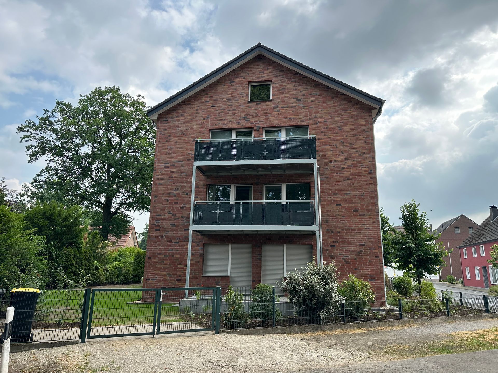 Backsteingebäude mit Balkonen, grünem Tor, bewölktem Himmel.