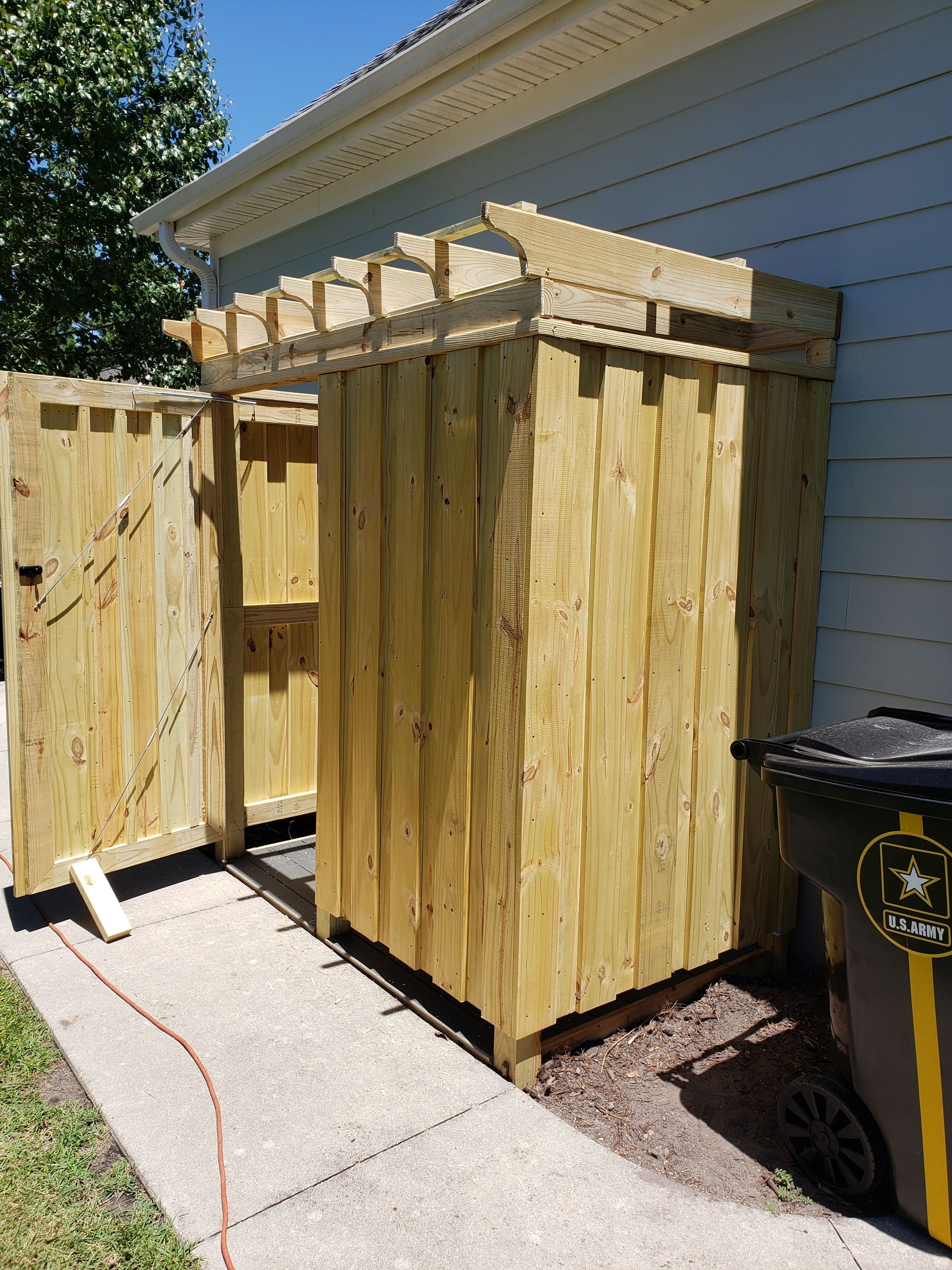 Wooden trash can enclosure with open door. Built against a light-colored house. Black trash can visible.