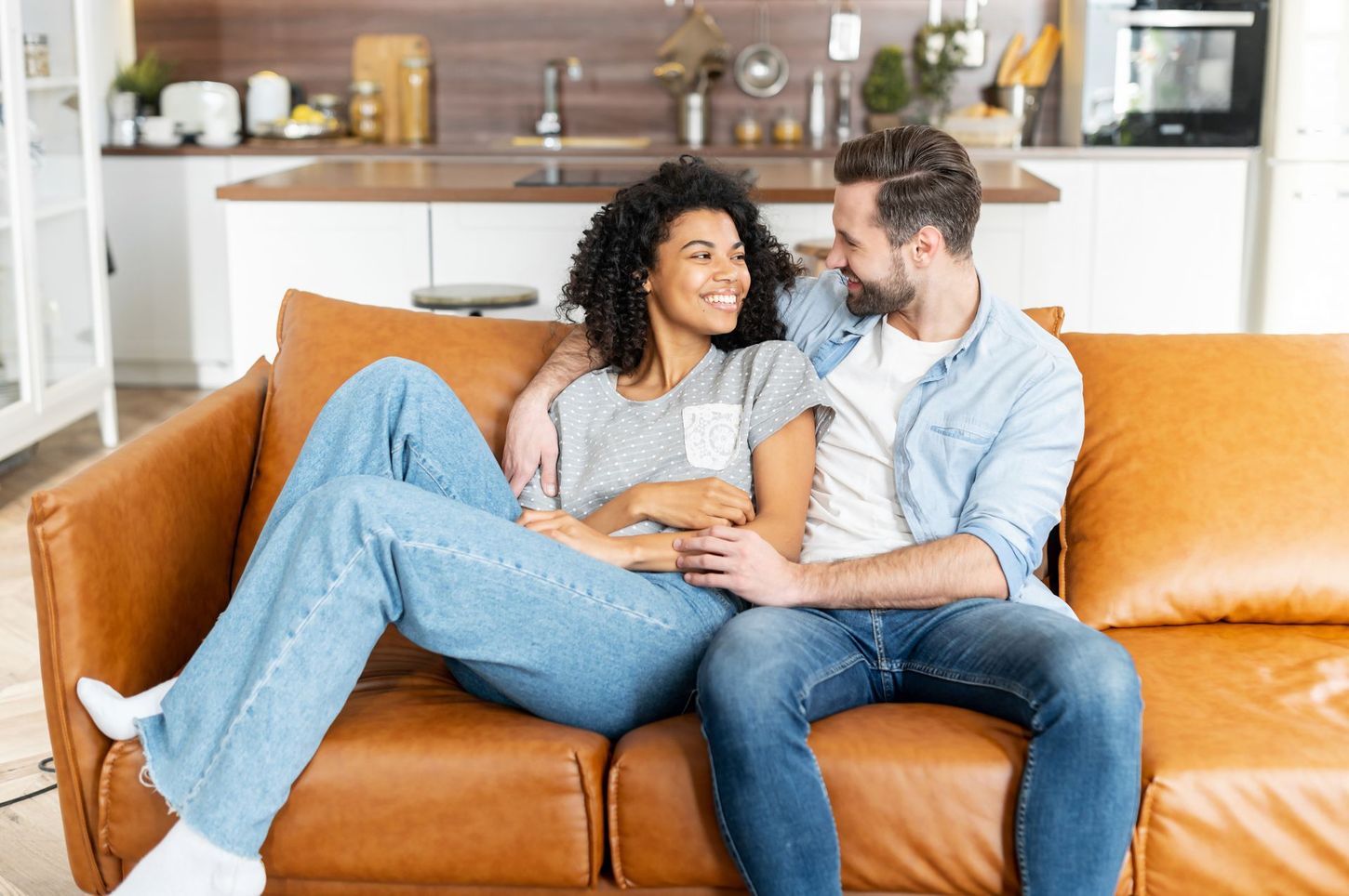 Couple sitting close together on a brown couch, smiling at each other. Kitchen background.
