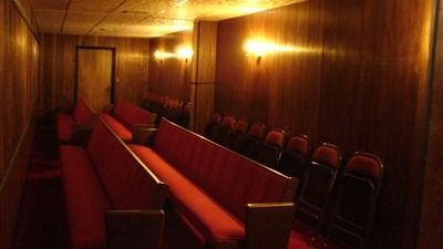 Empty, wood-paneled room with rows of red padded benches, lit by warm lights. A door is at the end.