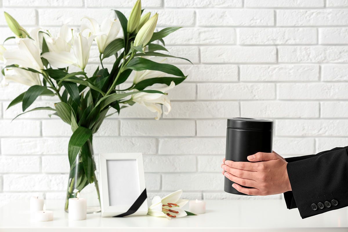 Person holding black urn, with white lilies and a framed photo on a white shelf against a white brick wall.